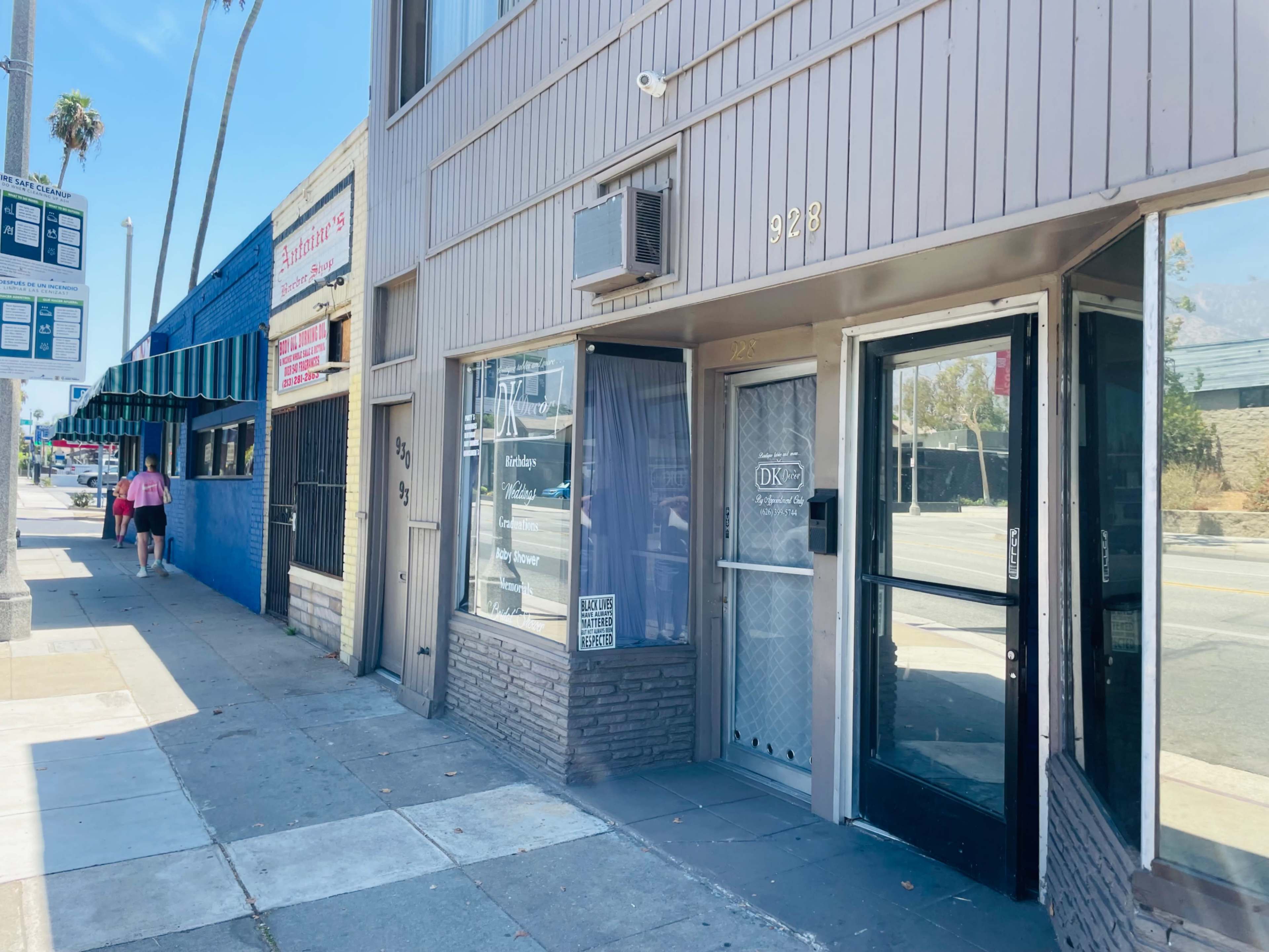 The image shows a storefront with a glass door and a sign on the window, located on a sidewalk near palm trees and other buildings.