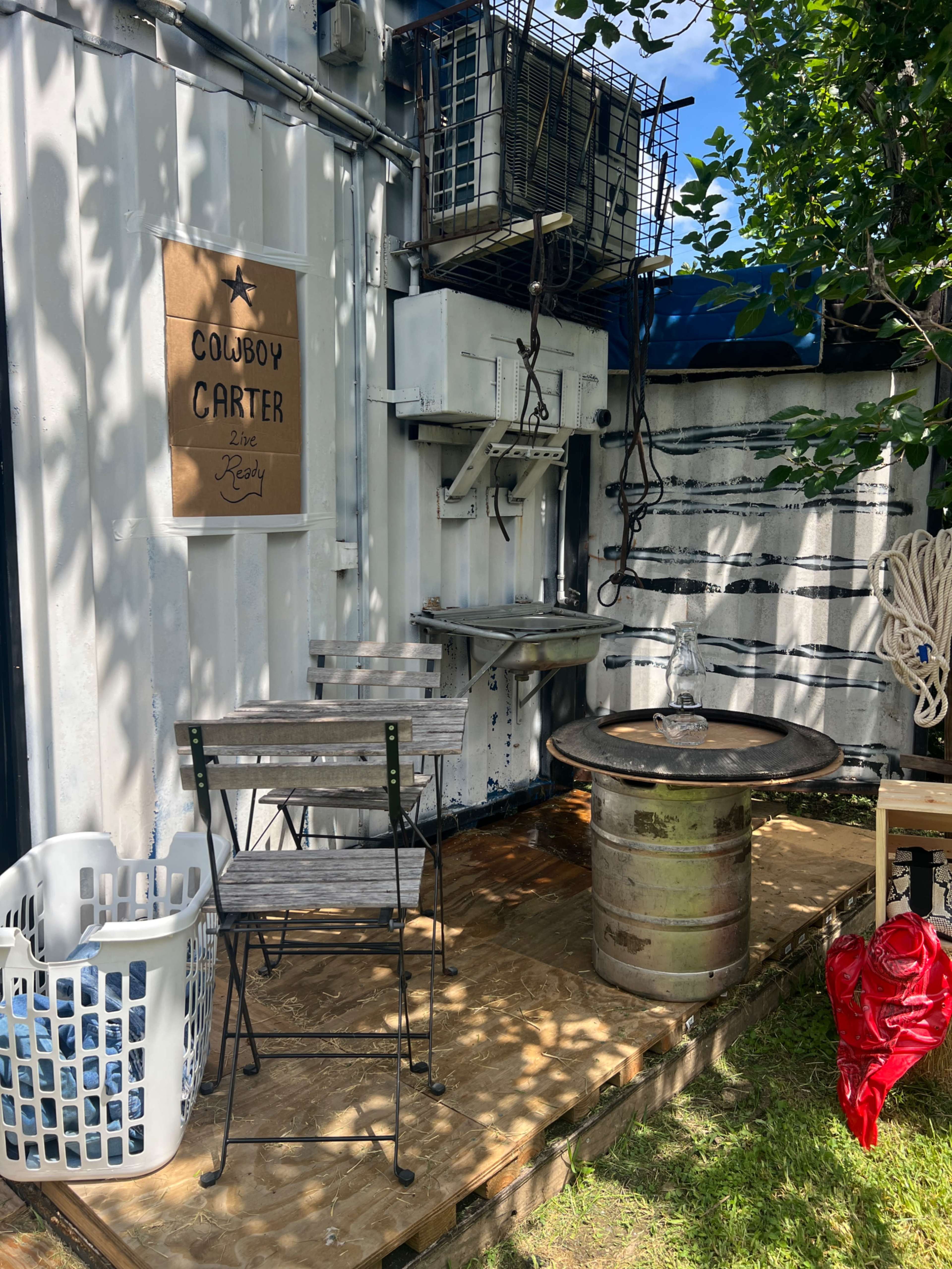 The image shows a small outdoor area featuring a chair, a table made from a metal drum, a laundry basket, and a sign that reads "COWBOY CARTER."