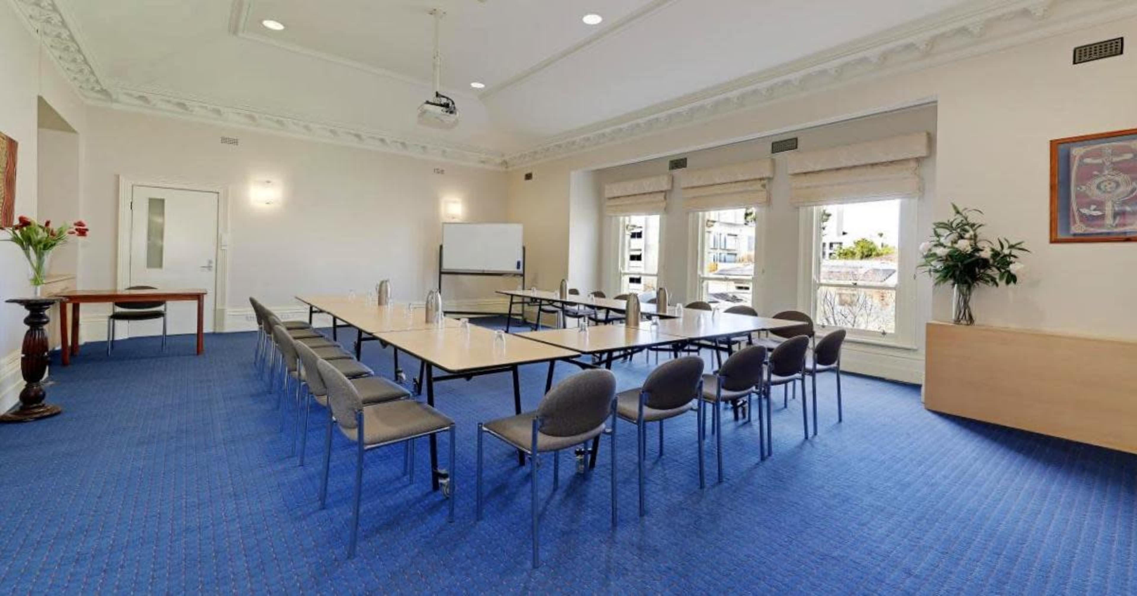 A conference room features a long table surrounded by chairs, with a projector and windows letting in natural light.