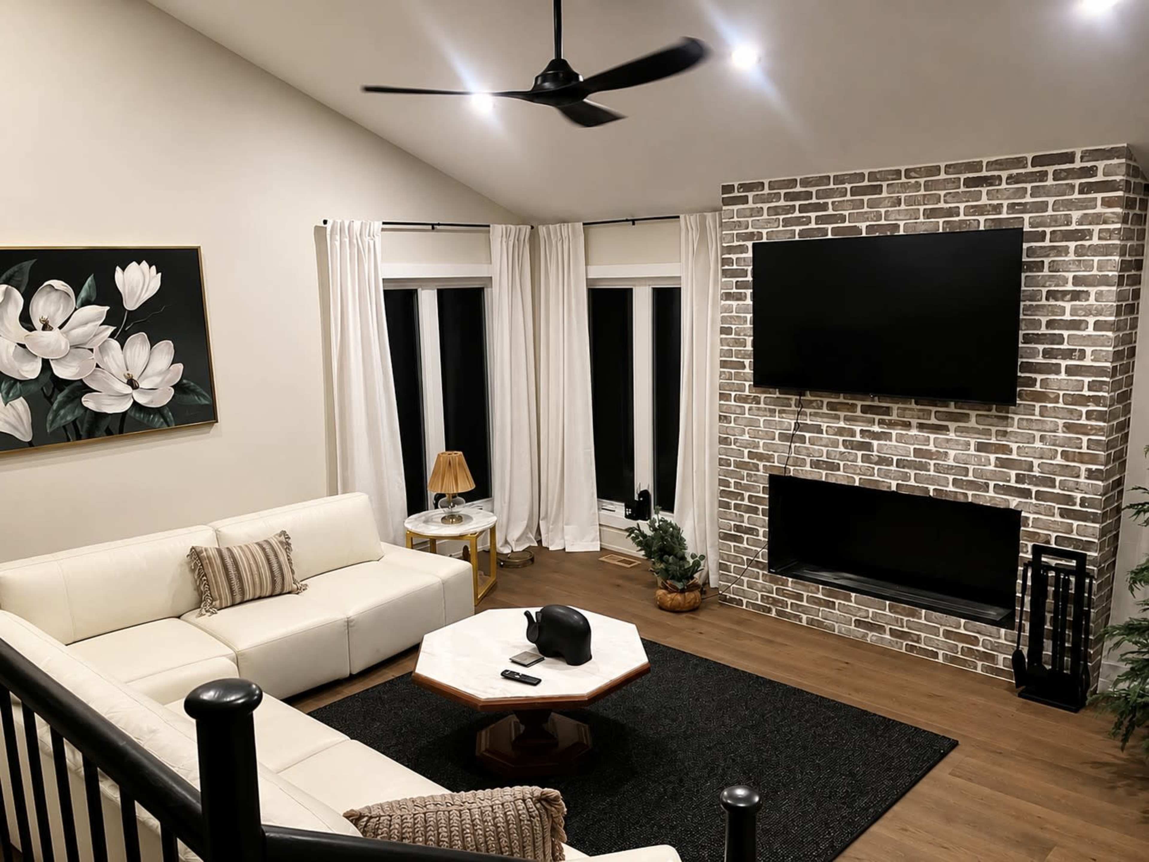 The image depicts a modern living room featuring a white leather sofa, a black area rug, a brick wall with two mounted flat-screen TVs, and large windows covered with white curtains.