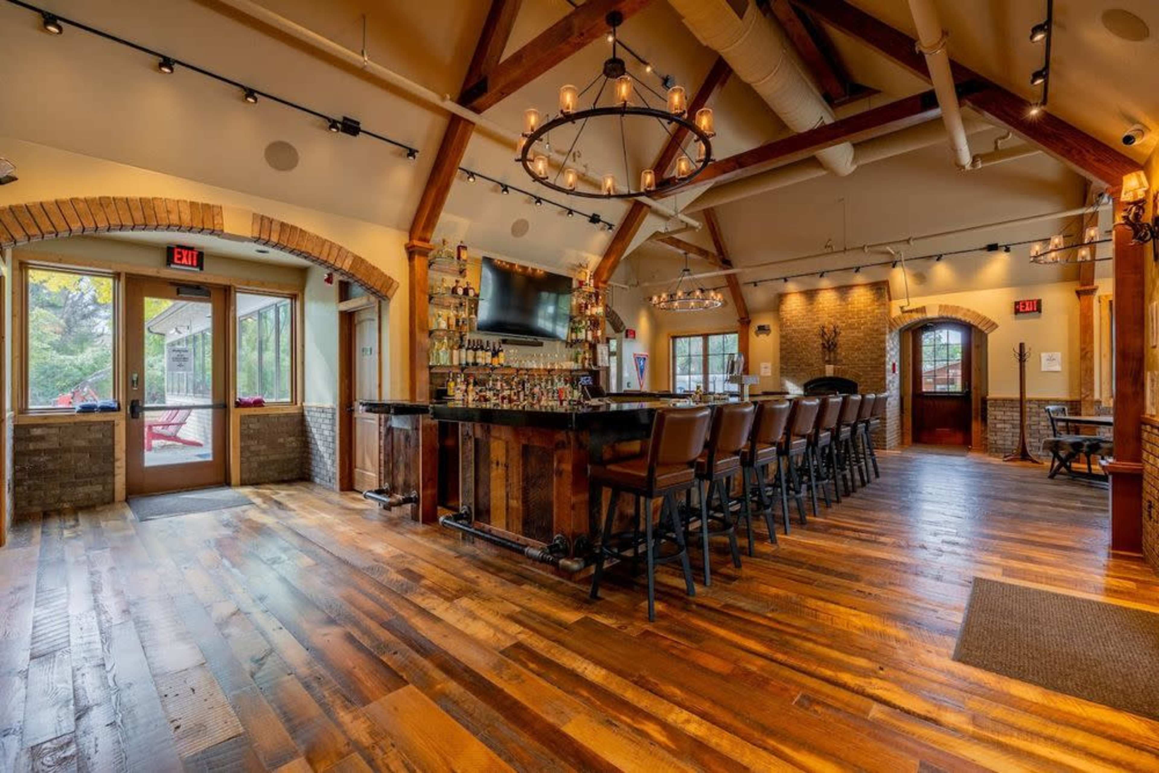 The image shows a rustic bar area with wooden floors, a long bar counter lined with high chairs, and large windows letting in natural light.