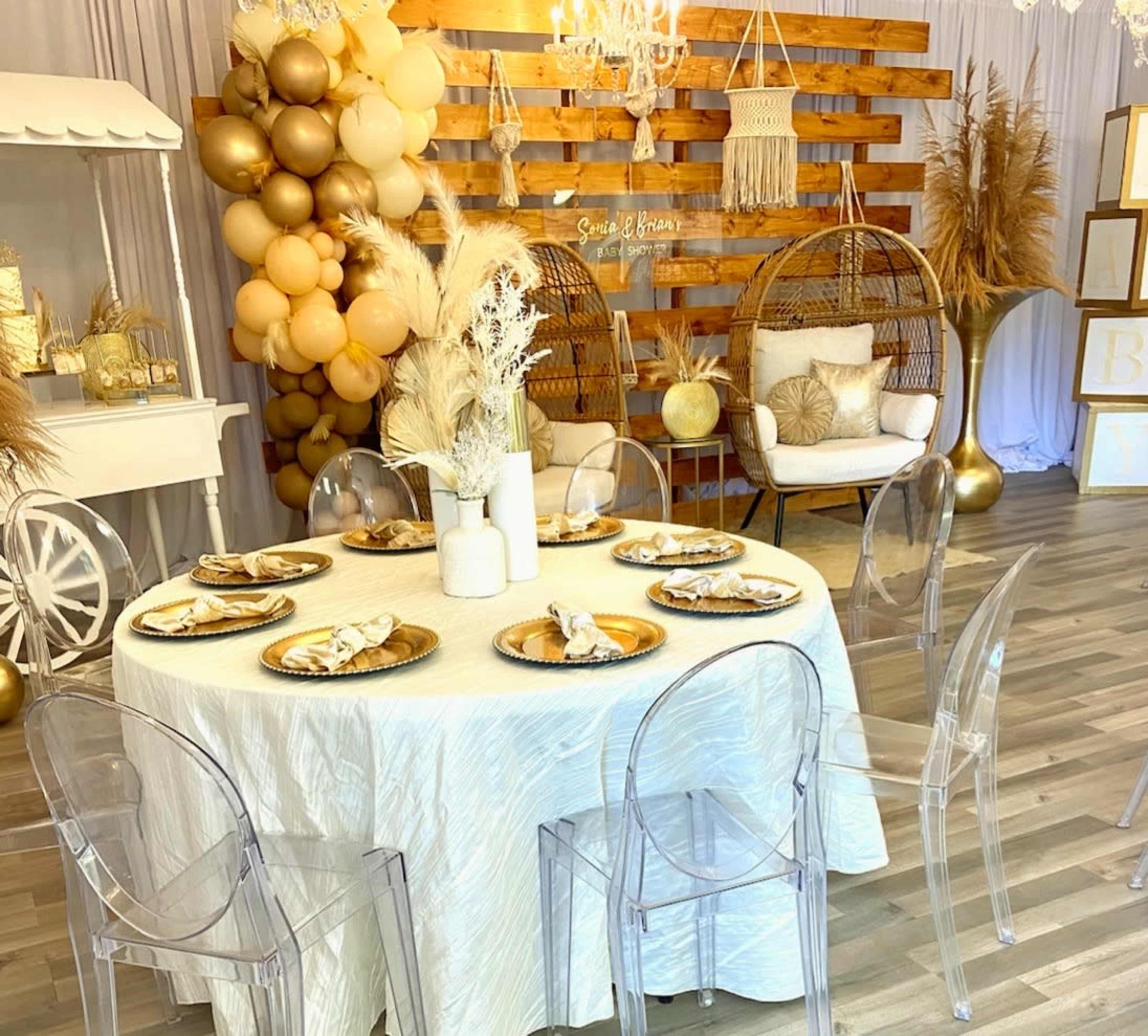 A decorated room with a round dining table set for a gathering, surrounded by clear acrylic chairs, and featuring balloons, a hanging chandelier, and a rustic wooden backdrop.