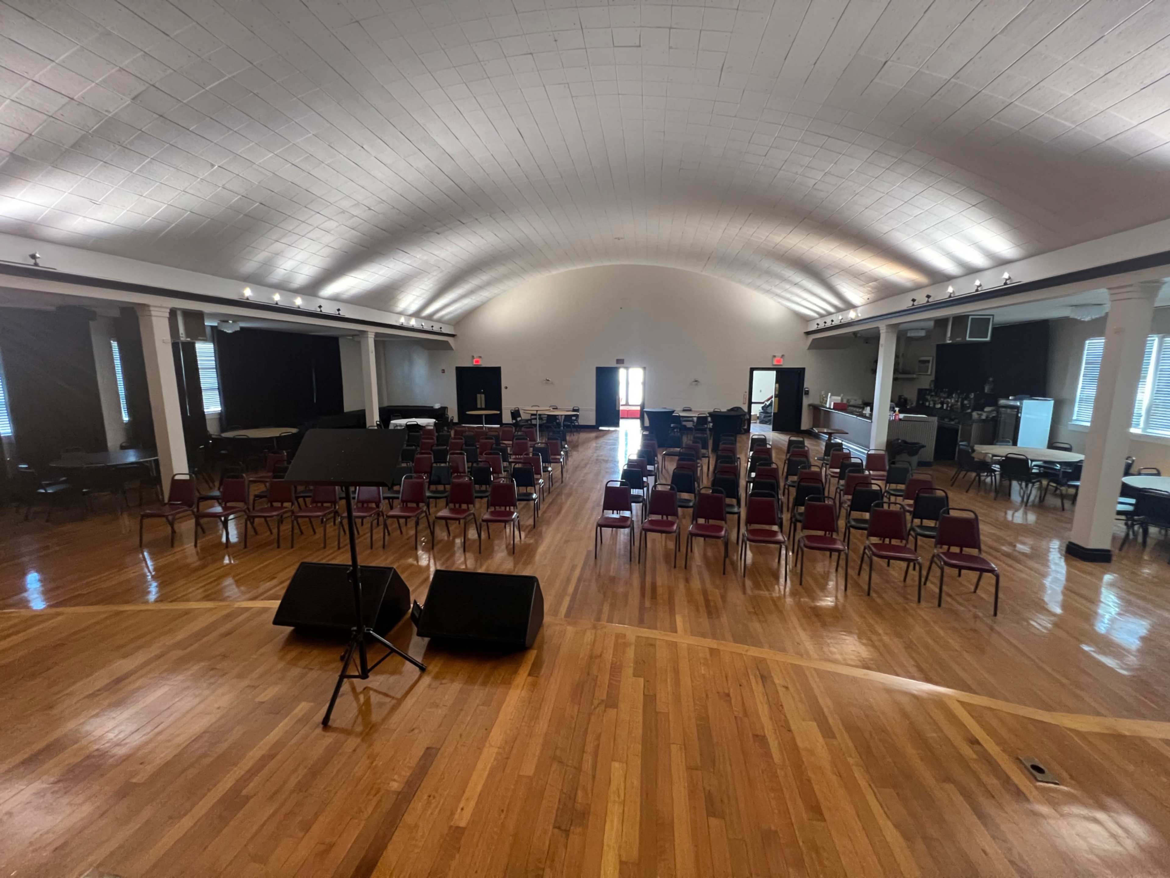The image shows a large, empty hall set up with rows of chairs and a stage, featuring wooden floors and a high ceiling.