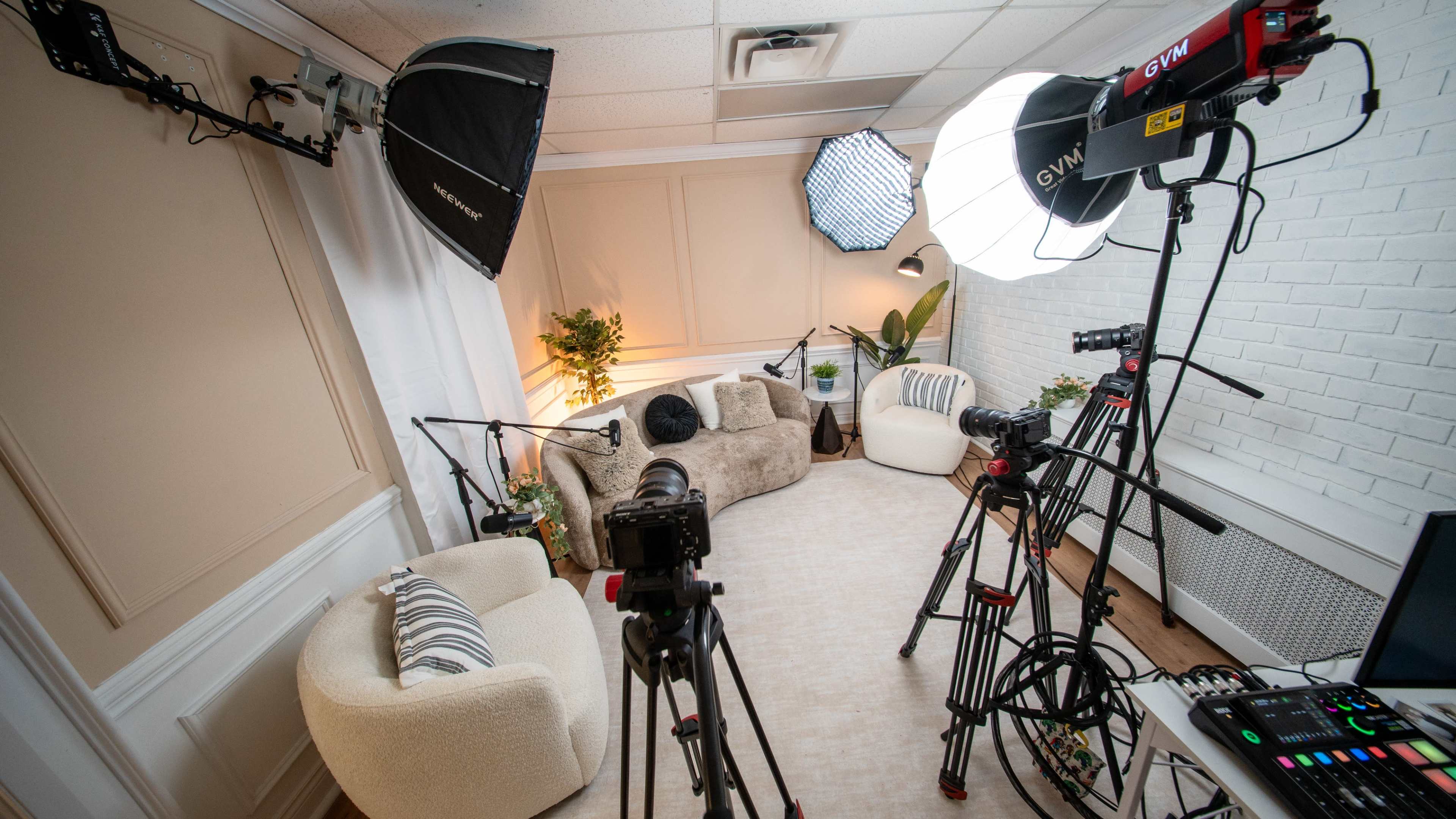 The image shows a video recording studio featuring three cameras, multiple softbox lights, and a cozy seating area surrounded by plants.