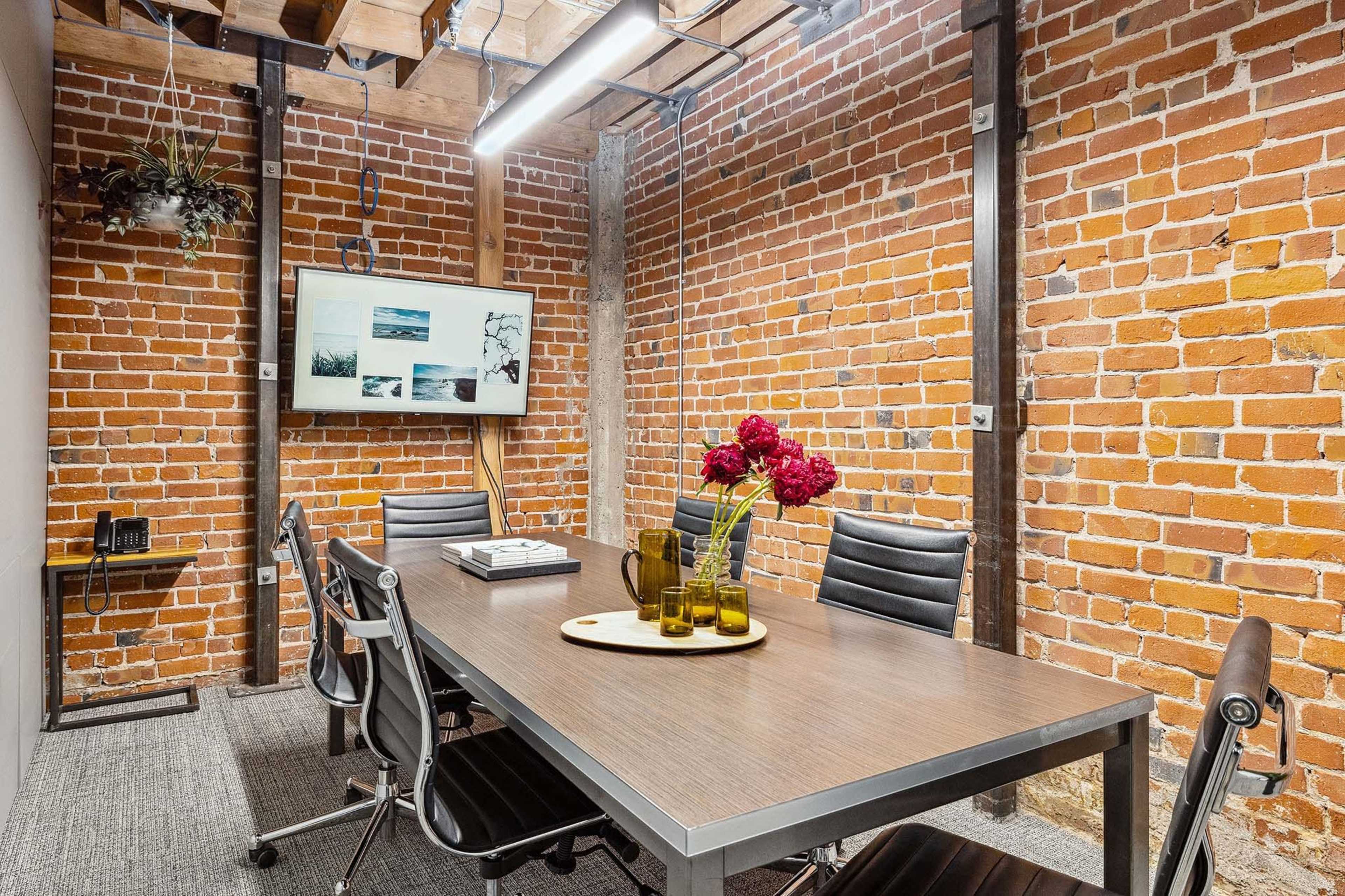 A conference room features a wooden table surrounded by black chairs, with exposed brick walls and a television displaying images.