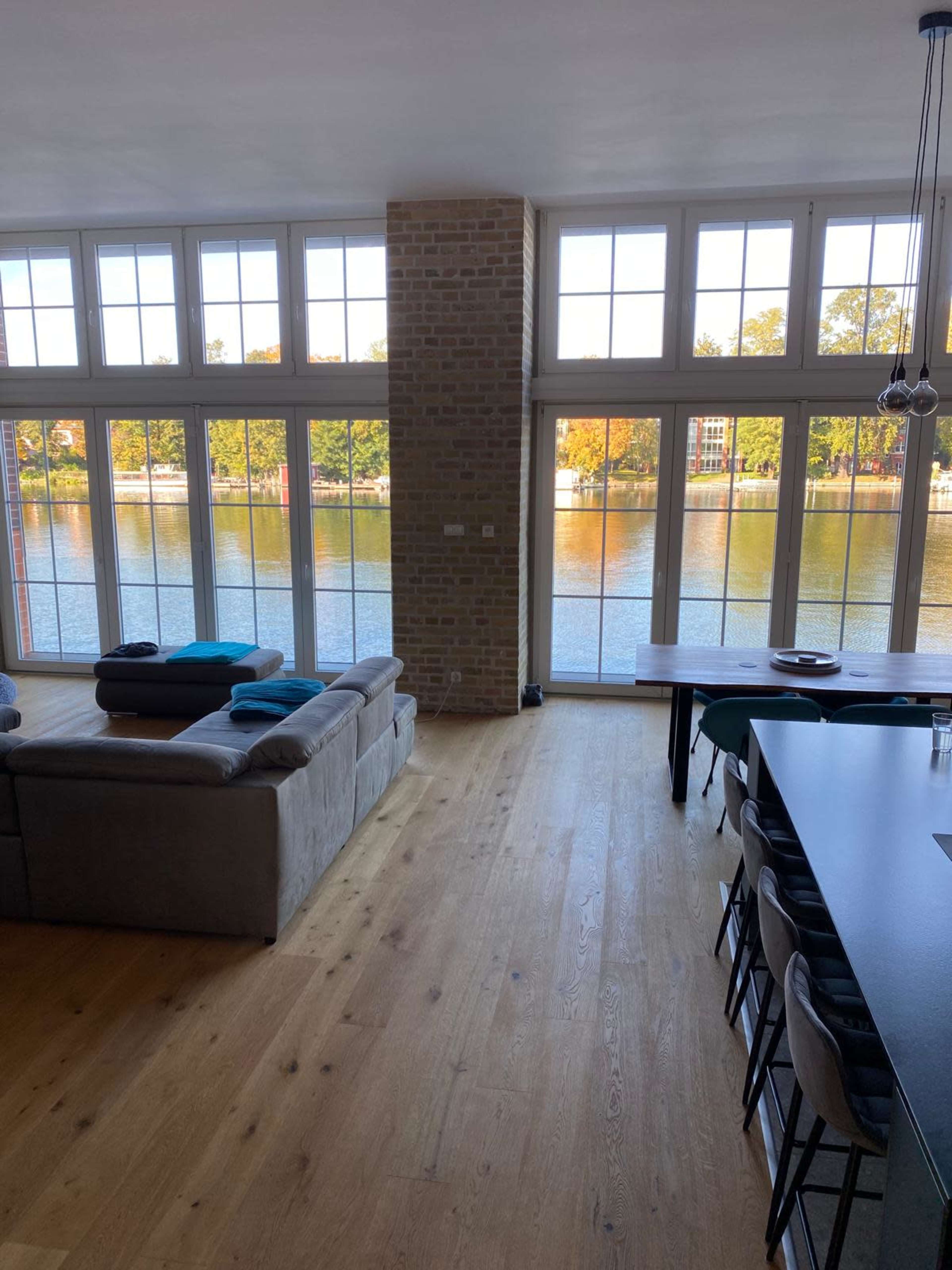 The image shows a bright, modern interior living space with large windows overlooking a body of water and a dining area with a long table.