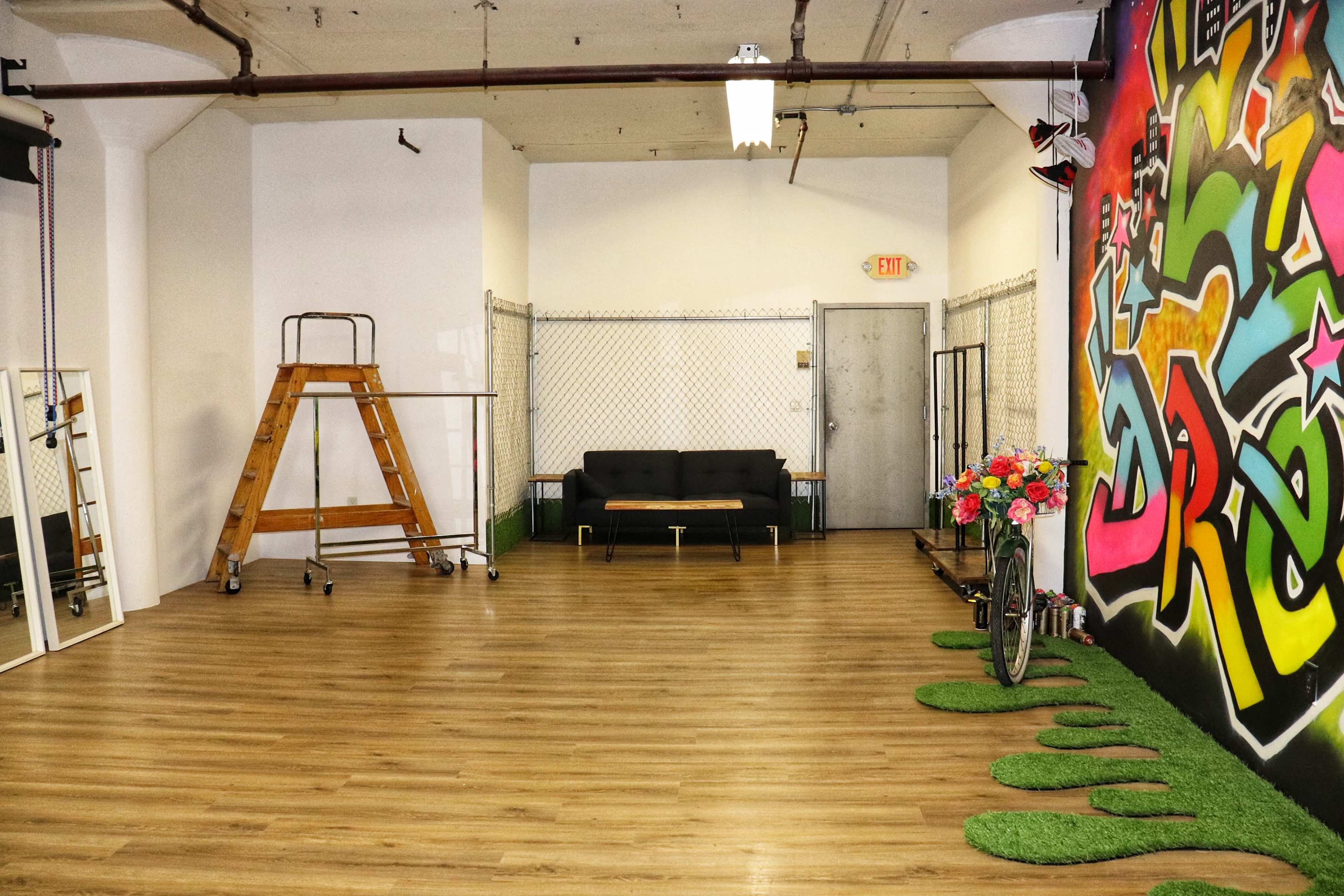 A spacious room with wooden flooring, a black couch against one wall, a colorful graffiti mural on another, and green grass-like decor on the floor.