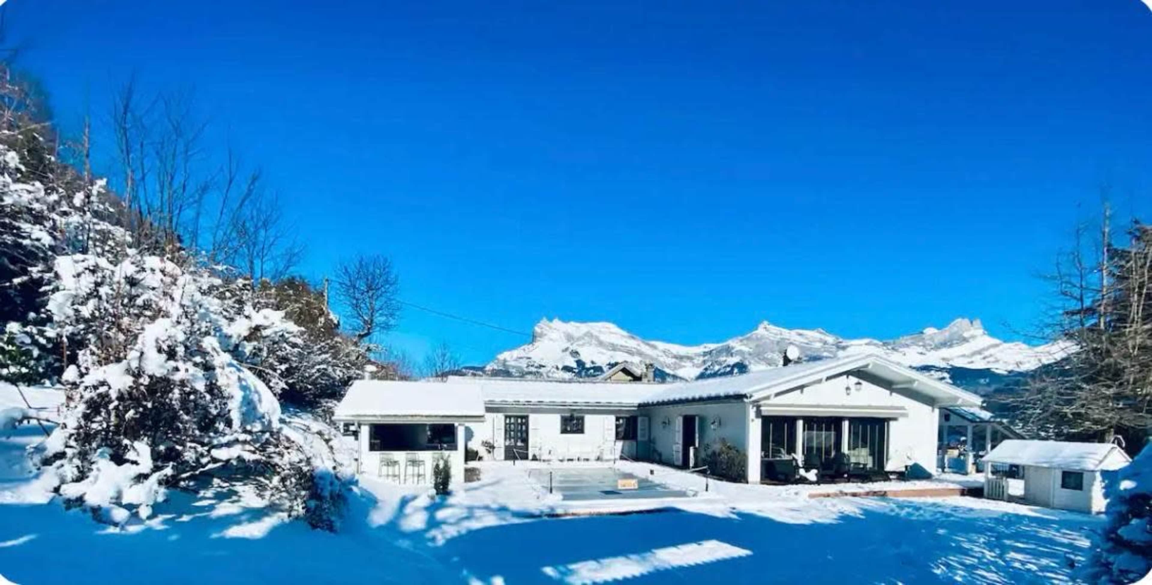 A snow-covered house sits against a backdrop of mountains under a clear blue sky.
