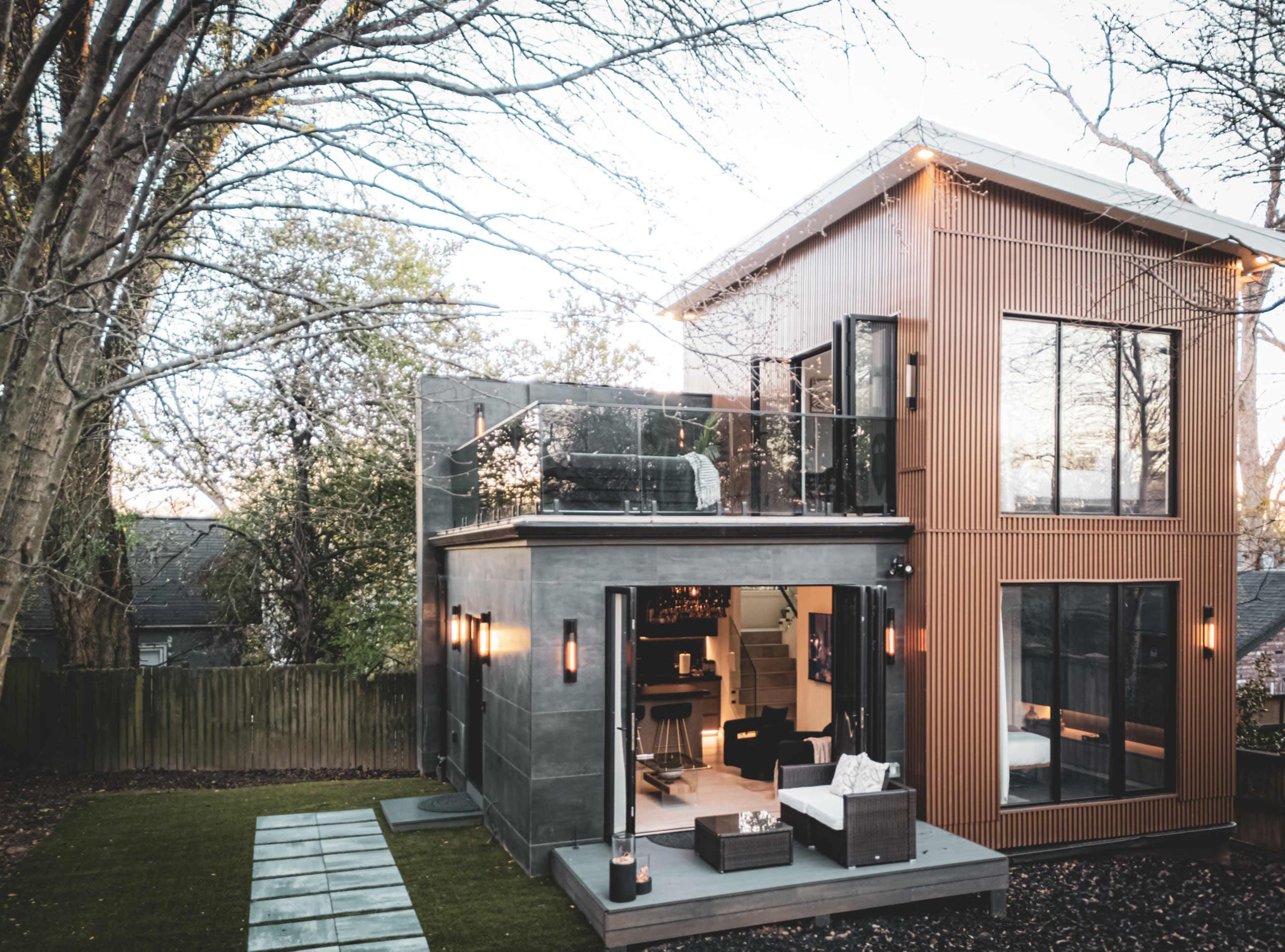 The image depicts a modern two-story house with a sleek exterior, featuring a glass terrace on the upper level and an open patio leading into a living space.