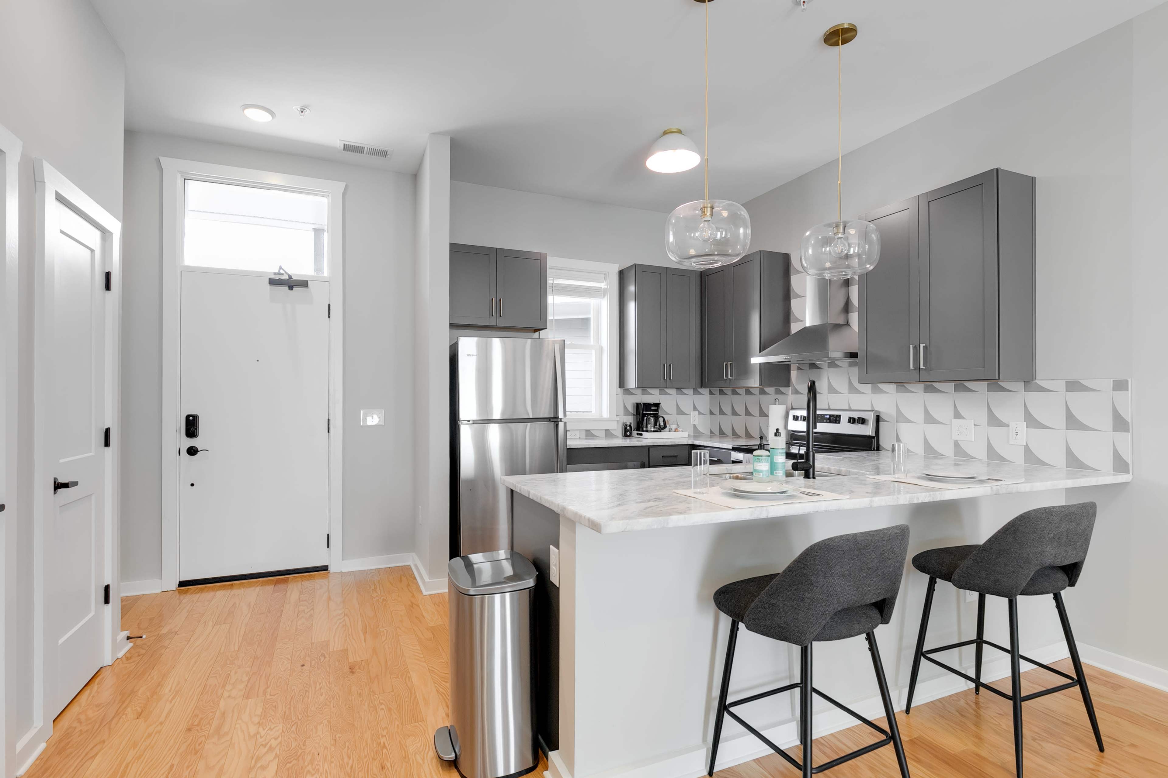 A modern kitchen with gray cabinetry, stainless steel appliances, and a breakfast bar with two stools, adjacent to a front door.