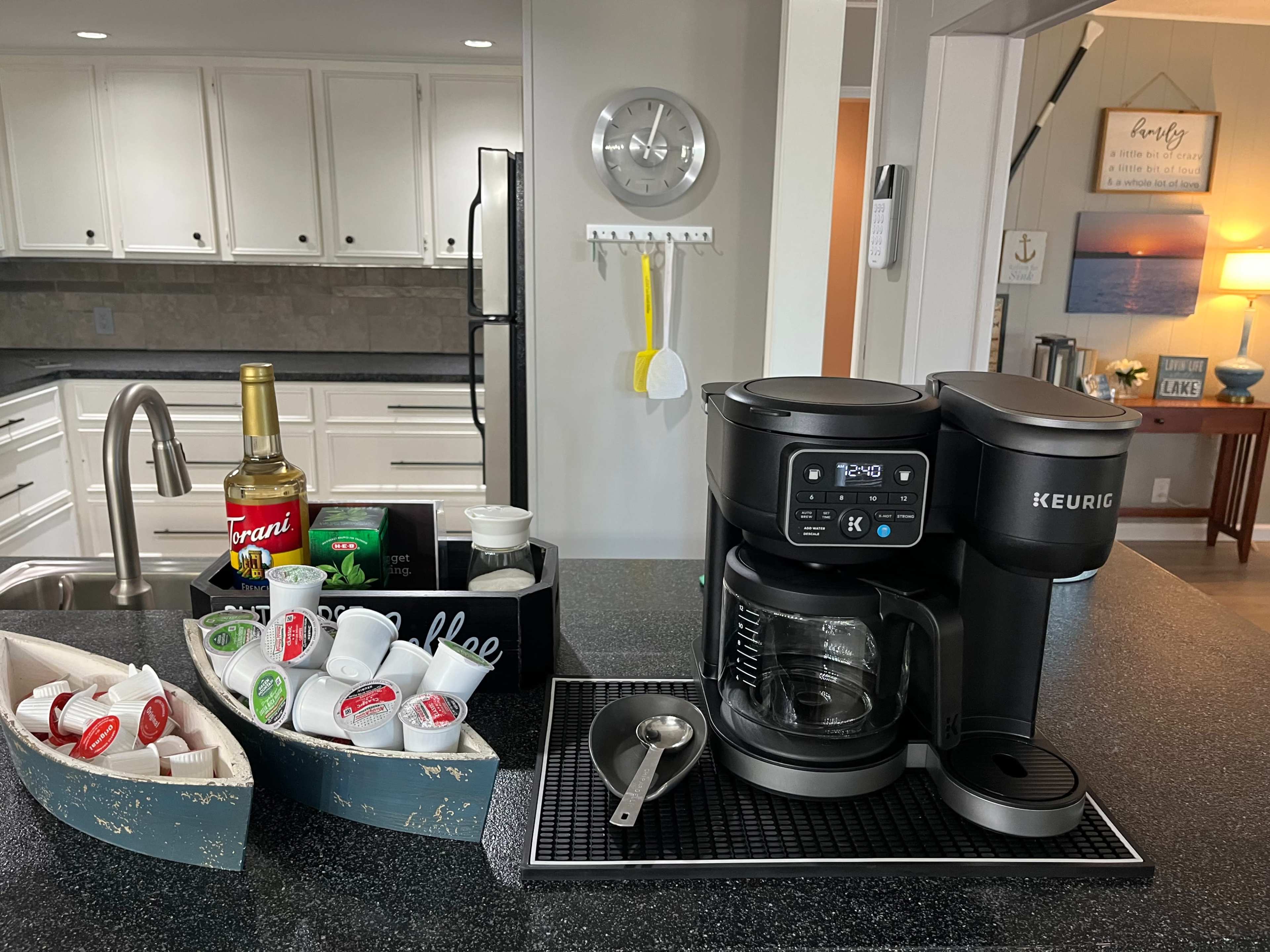 A Keurig coffee maker is positioned next to a countertop filled with coffee pods, creamers, a spoon, and other brewing accessories in a modern kitchen.