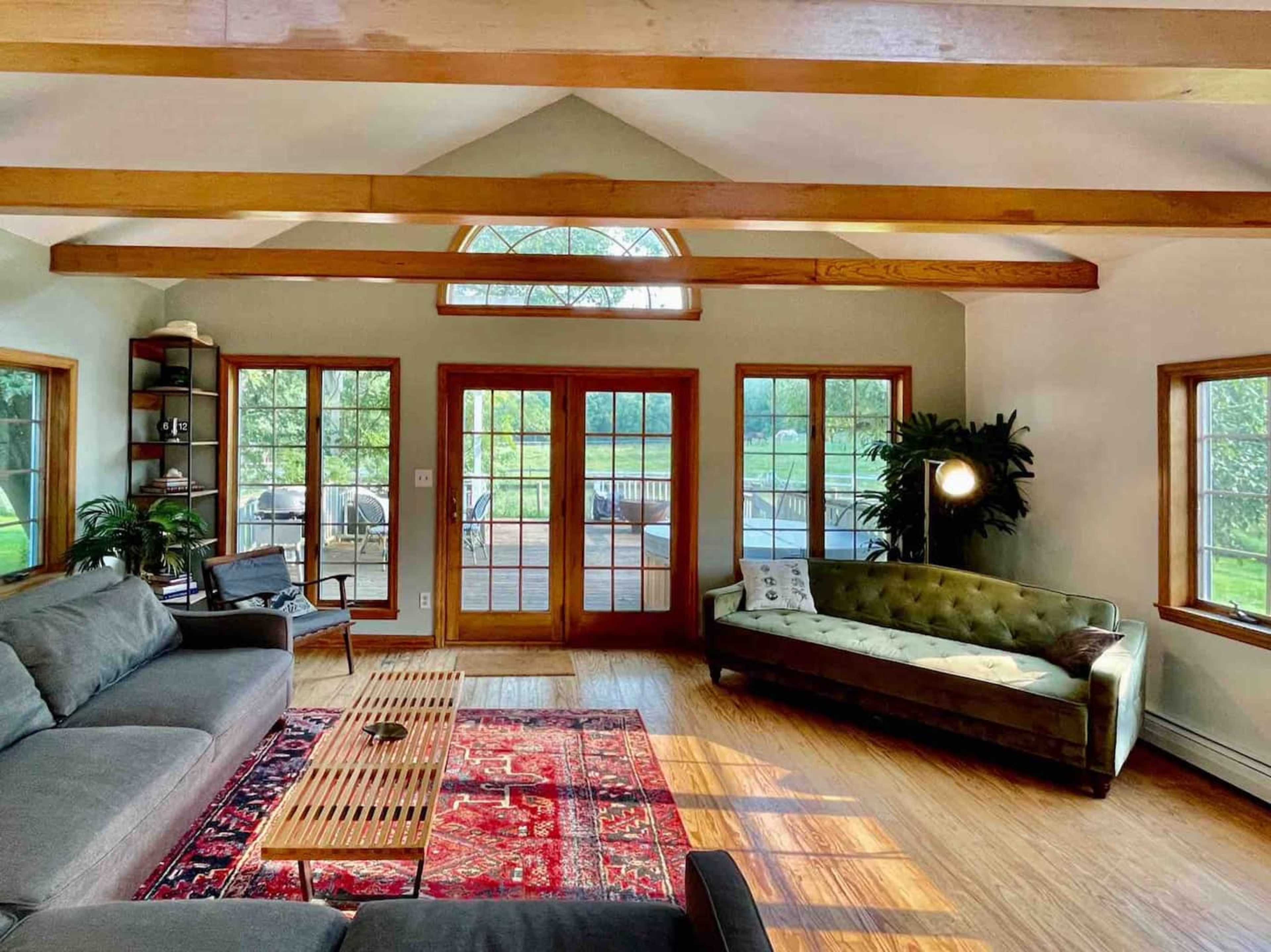 A bright living room with wooden beams, large windows, a gray sofa, a green tufted couch, a coffee table, and a patterned rug.