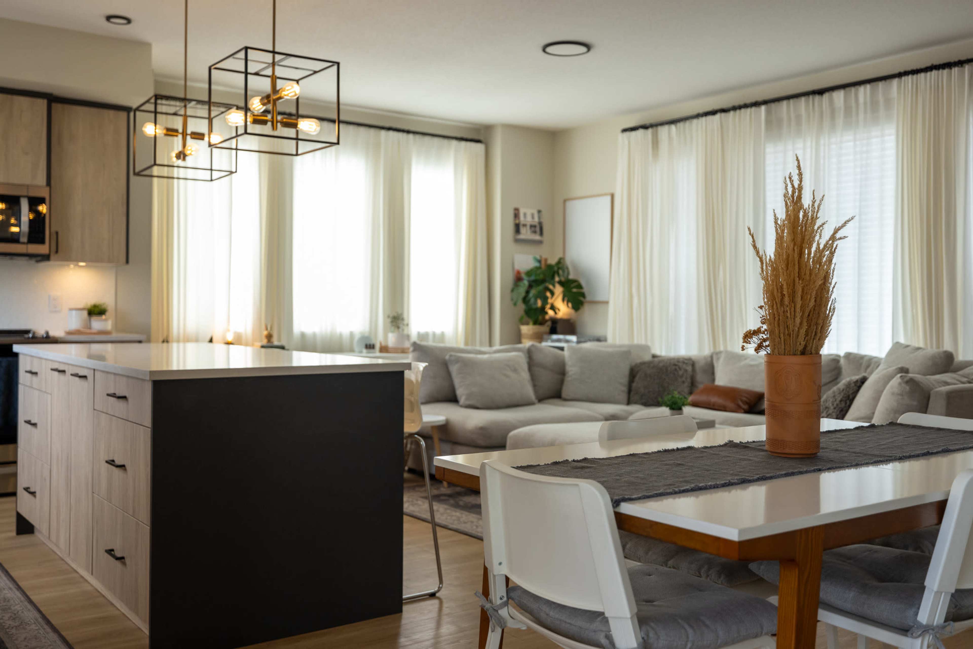 A modern kitchen and living area featuring a central island, a dining table, and large windows with sheer curtains.
