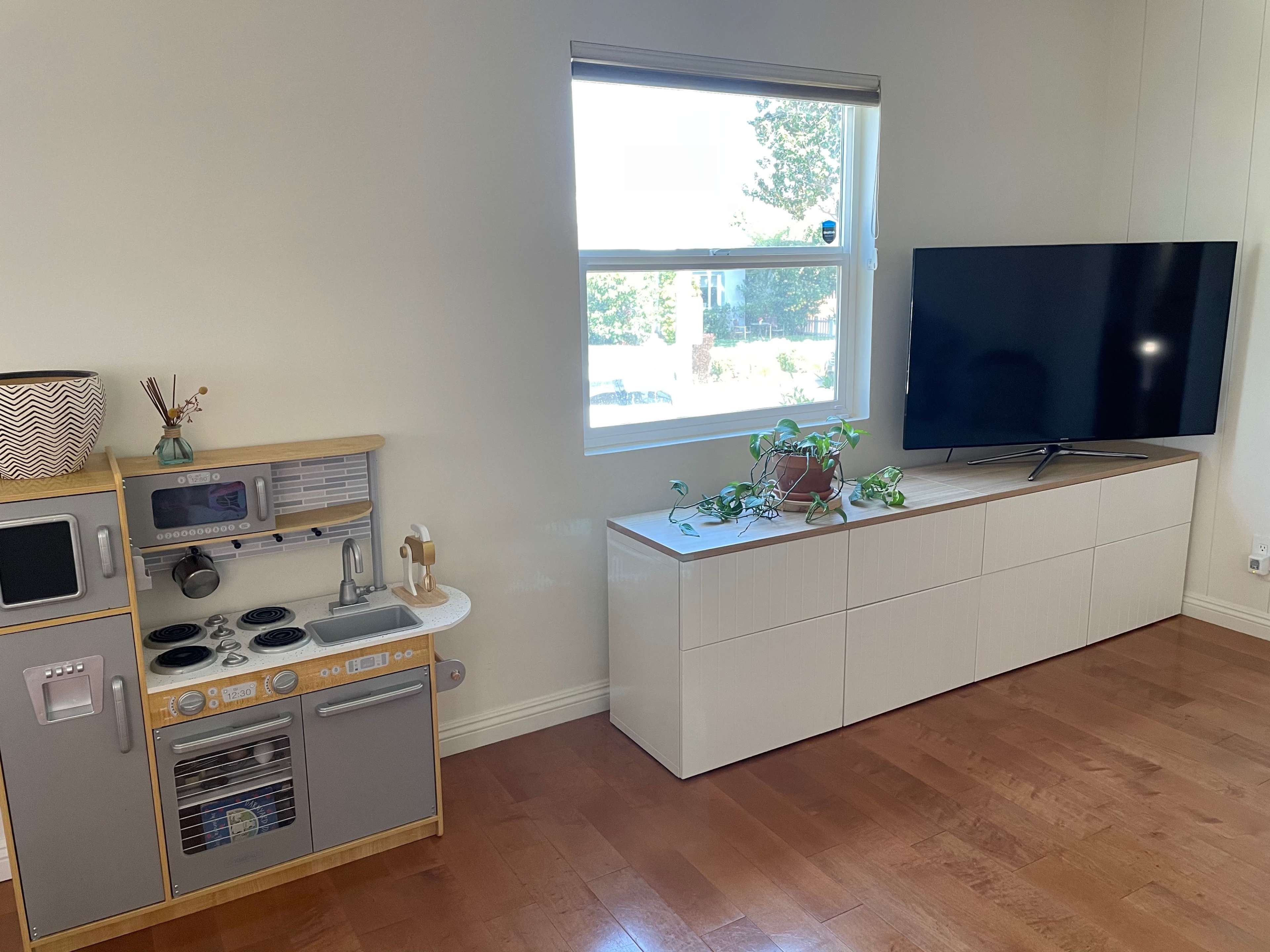 A children's play kitchen set is positioned next to a modern TV stand with a large flat-screen television in a well-lit room.