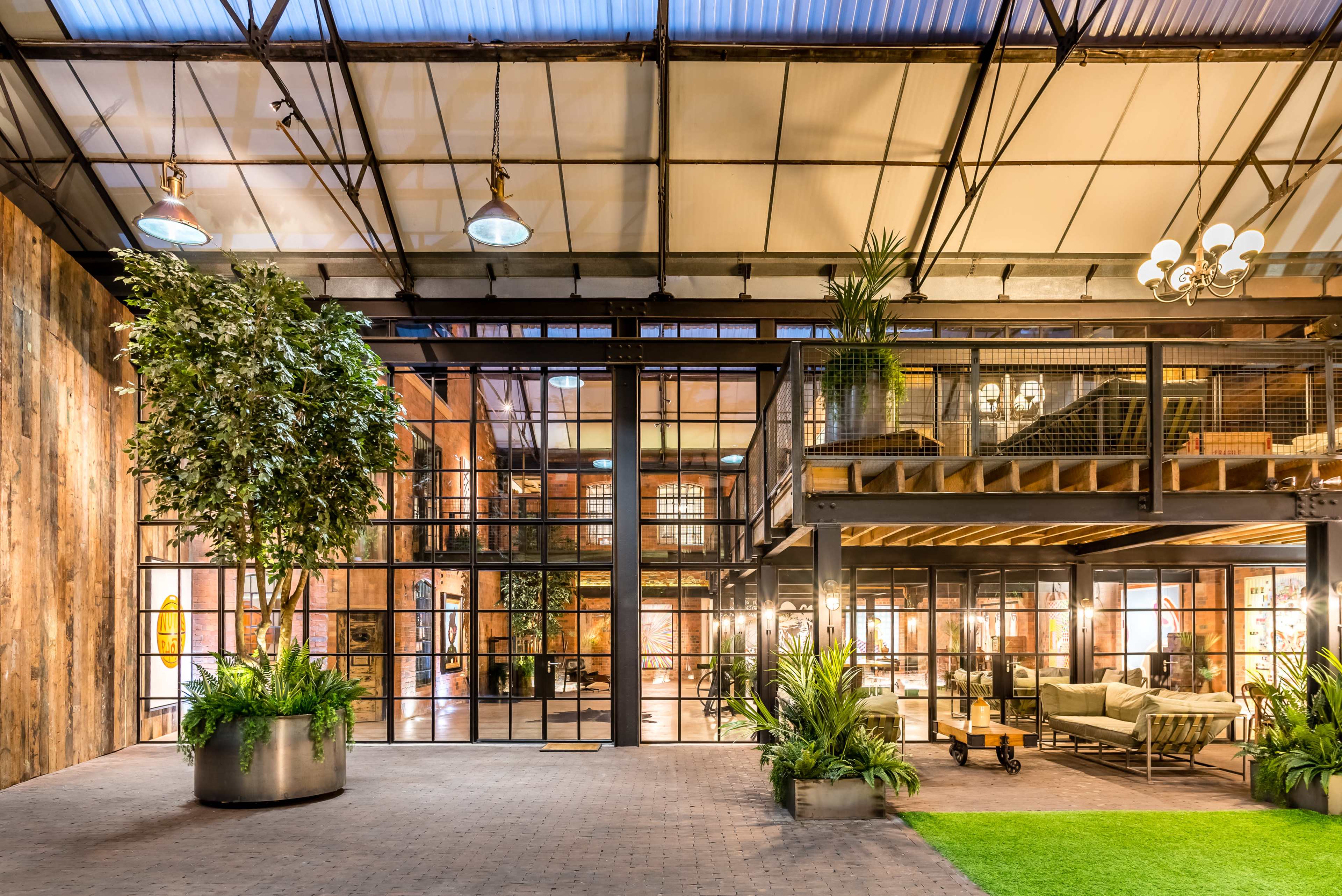 An expansive industrial-style interior with large glass windows, a mezzanine level, and greenery integrated into the space.