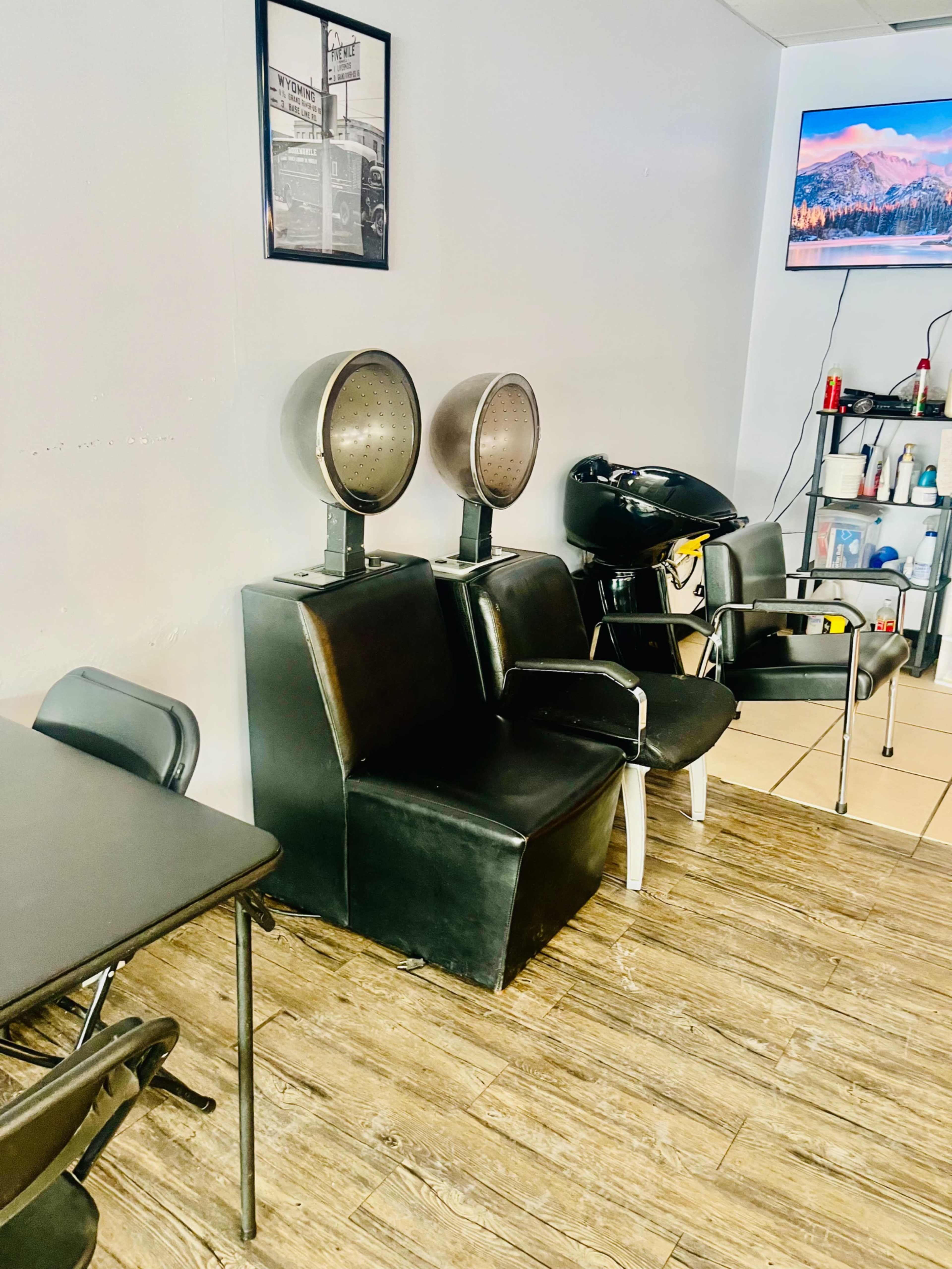 The image shows a salon interior with two hair dryers on stands, a black chair, a table, and a wall-mounted television.