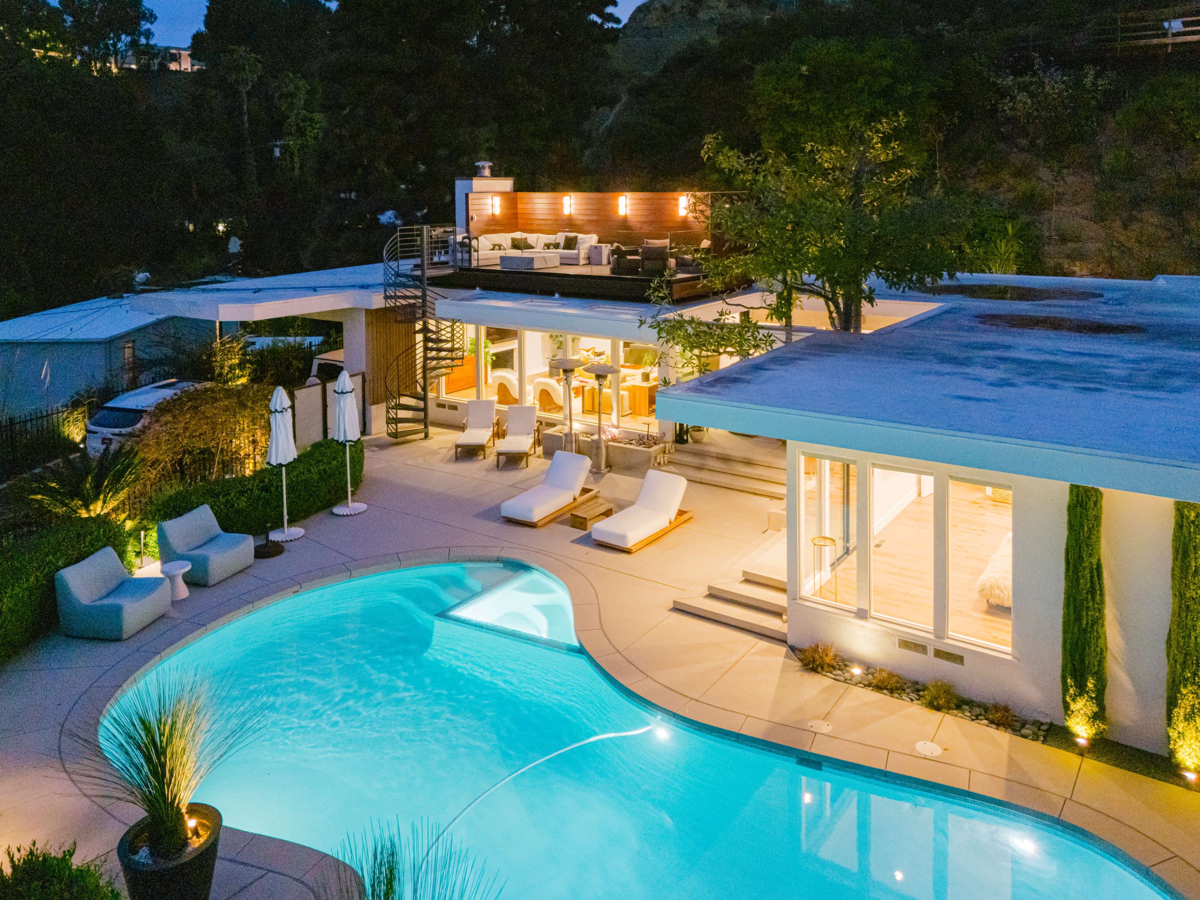 A modern villa features a lit swimming pool and a rooftop deck with seating, surrounded by landscaped greenery.