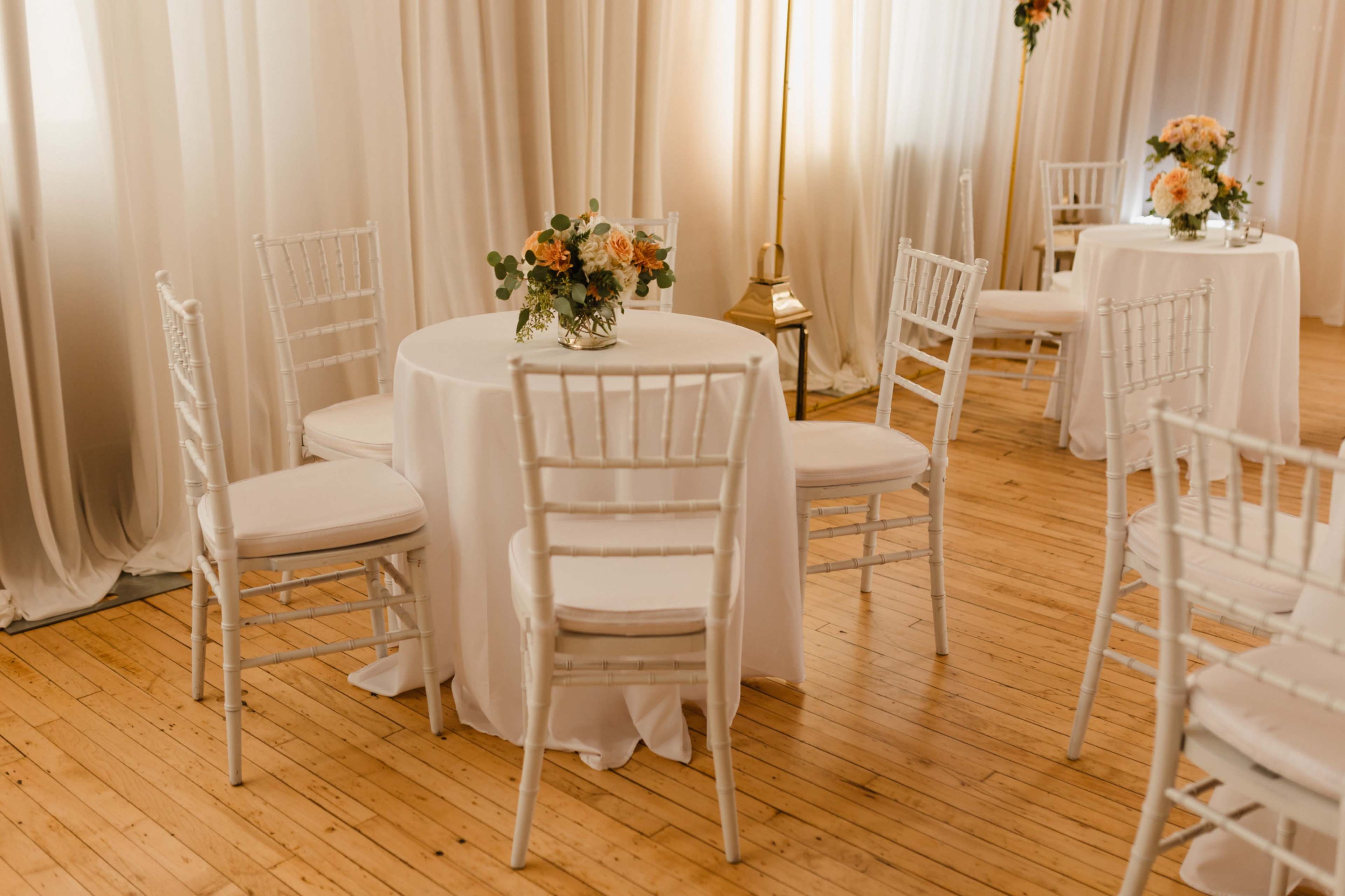 The image shows an elegantly arranged indoor event space with white tables, chiavari chairs, and floral centerpieces on each table.