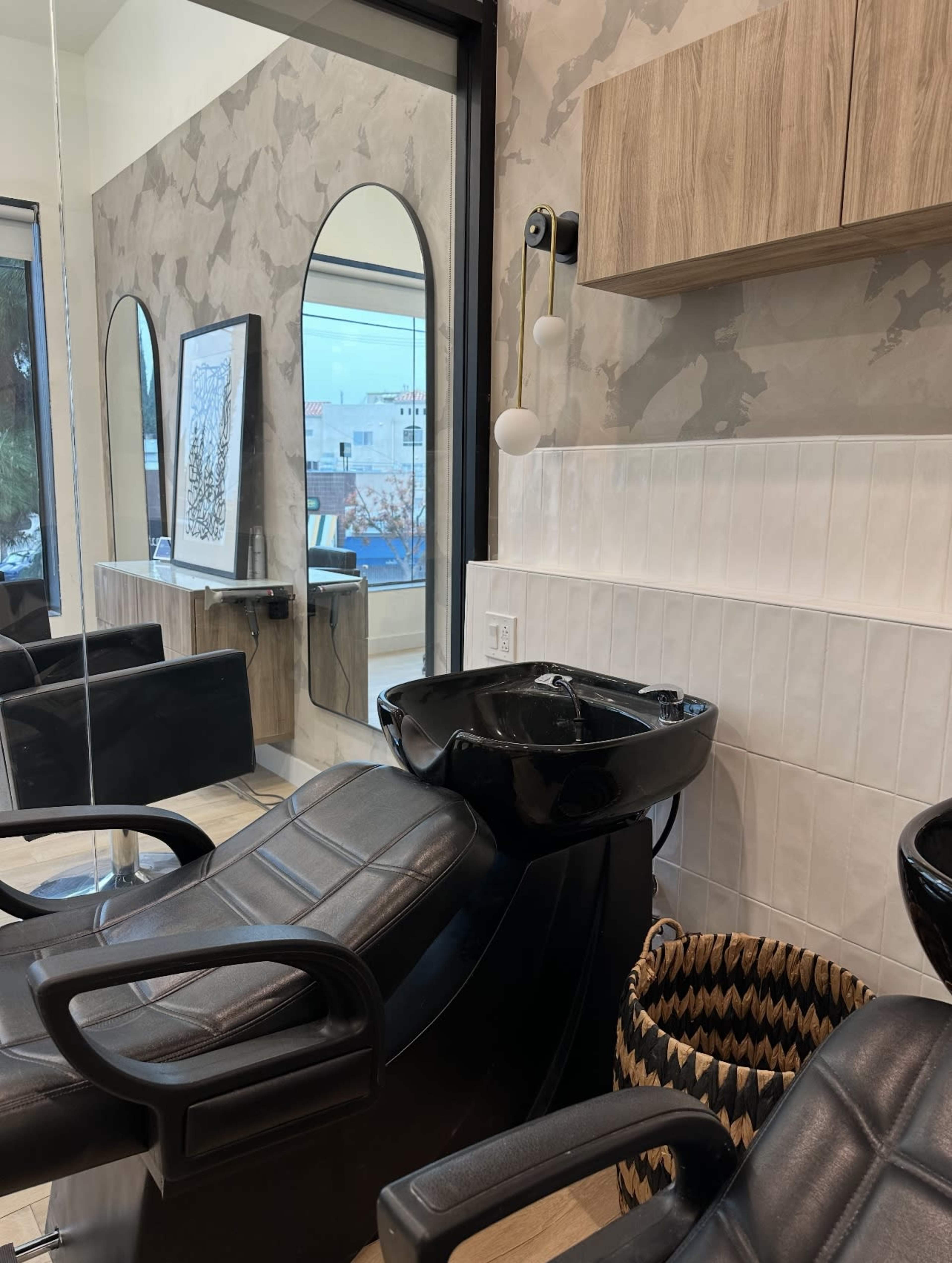 The image depicts a modern hair salon interior featuring dark leather shampoo chairs, a wall-mounted mirror, and a washing station with a black sink.