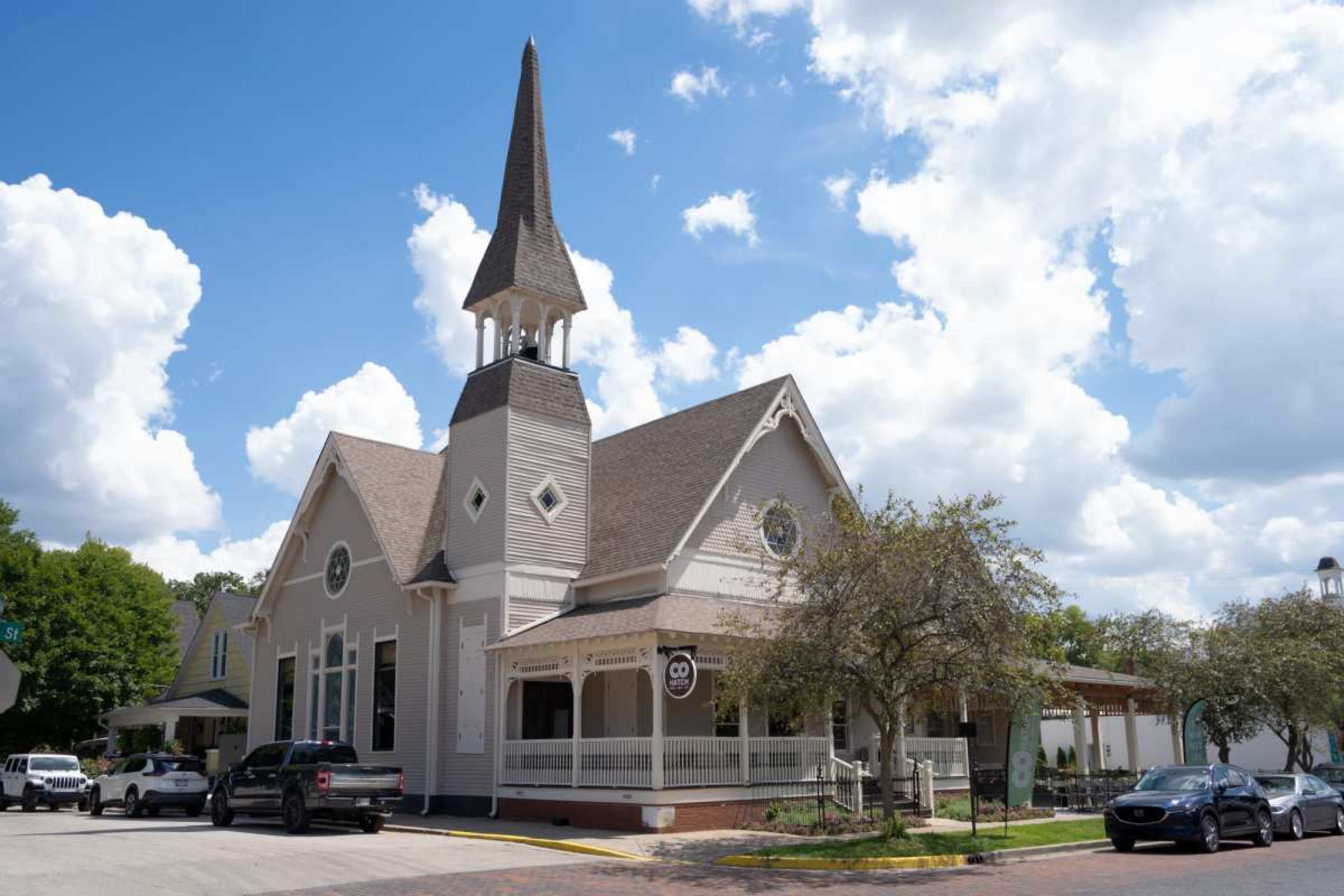 A large, white church with a tall steeple and a covered porch is seen against a blue sky filled with fluffy clouds.