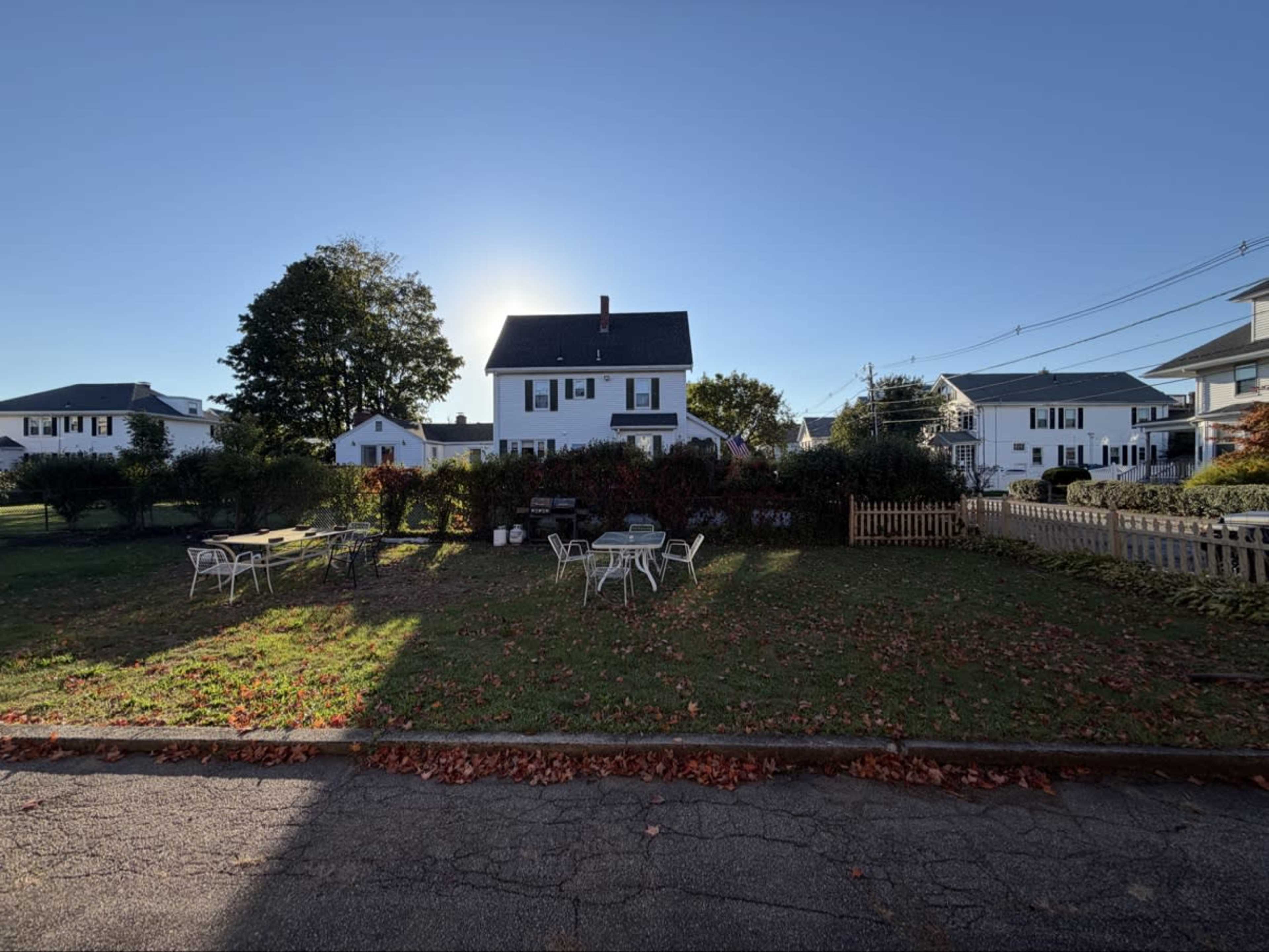 A white house with a green roof stands behind a yard scattered with fallen leaves, featuring several chairs and tables.