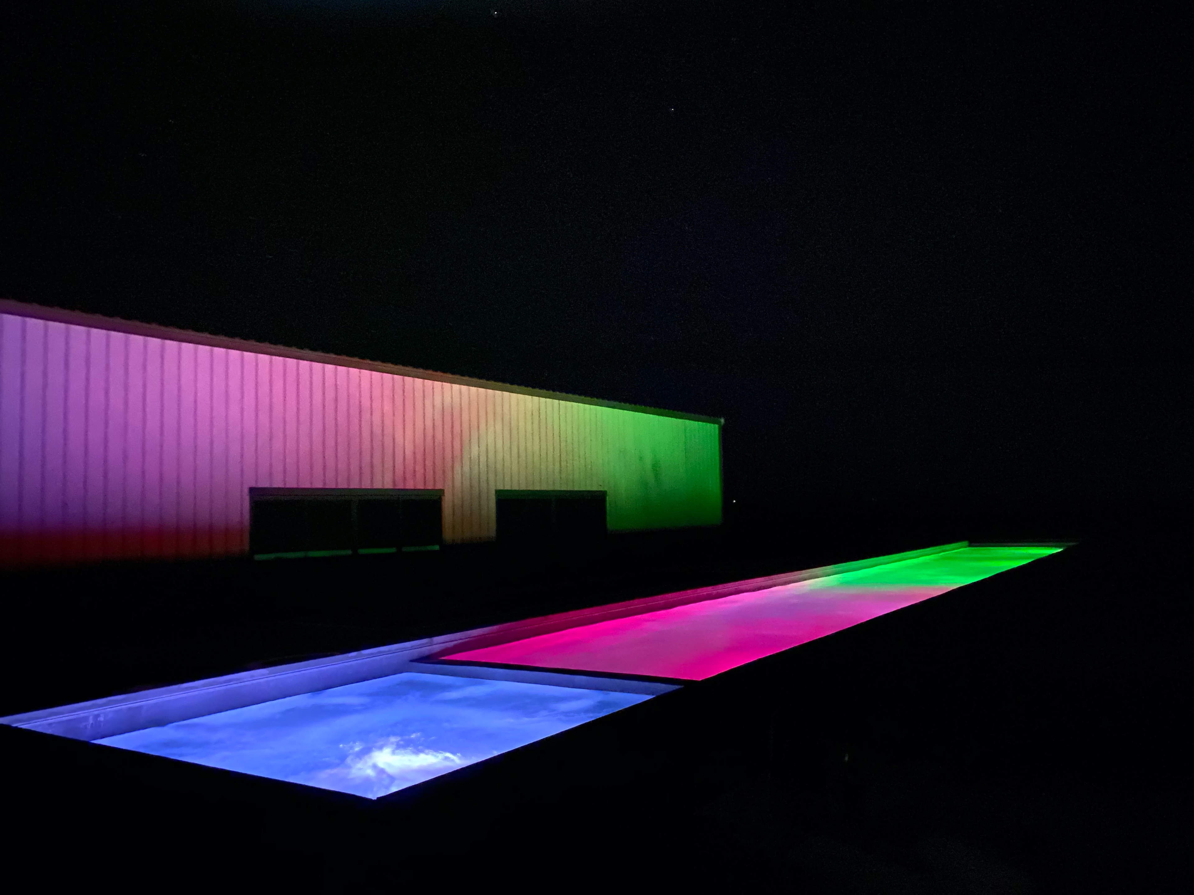 A long, illuminated outdoor pool is flanked by a building with colorful lighting in the background at night.