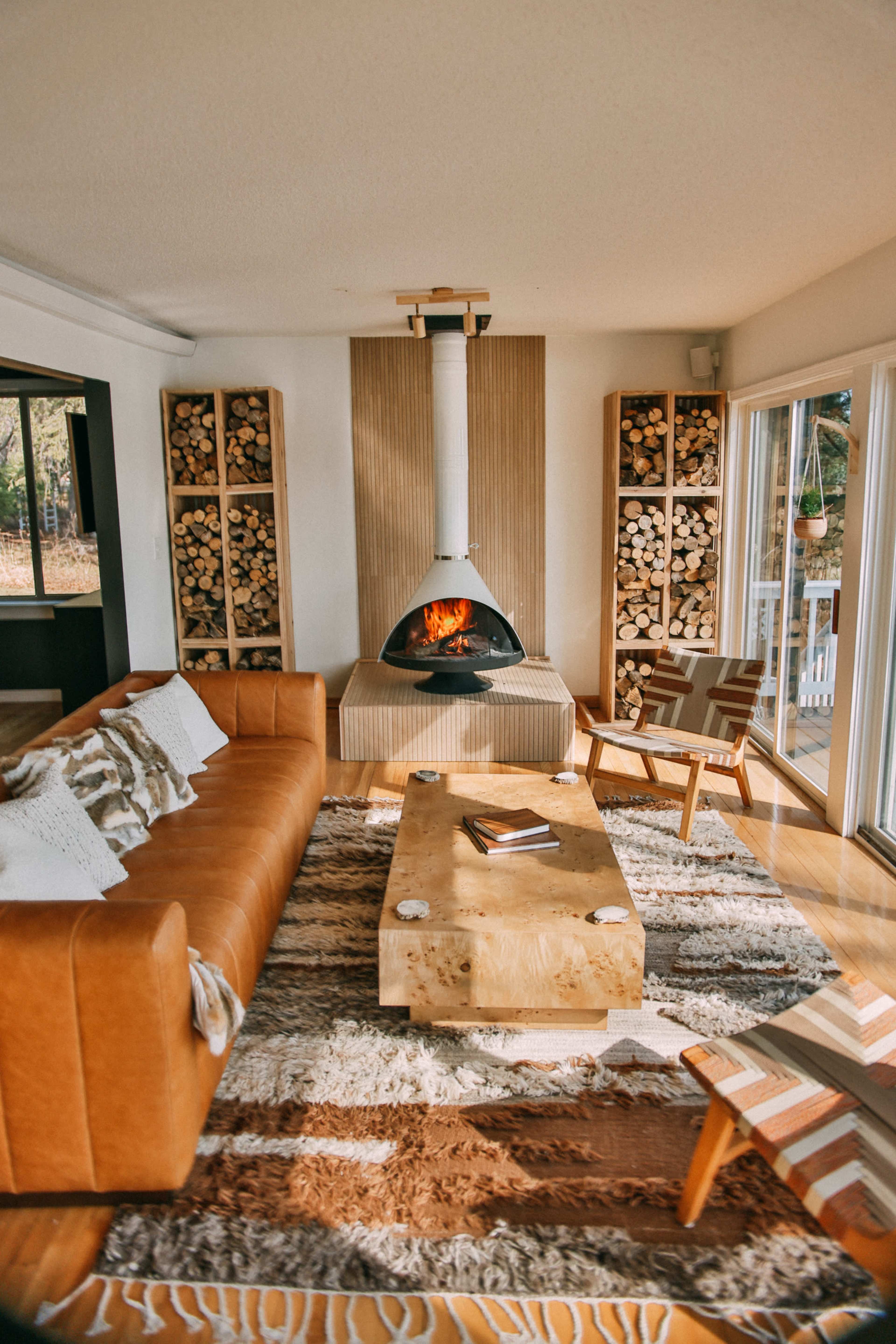 The image shows a modern living room featuring a round fireplace in the center, surrounded by wooden shelves stacked with logs, a leather sofa, a wooden coffee table, and large windows letting in natural light.