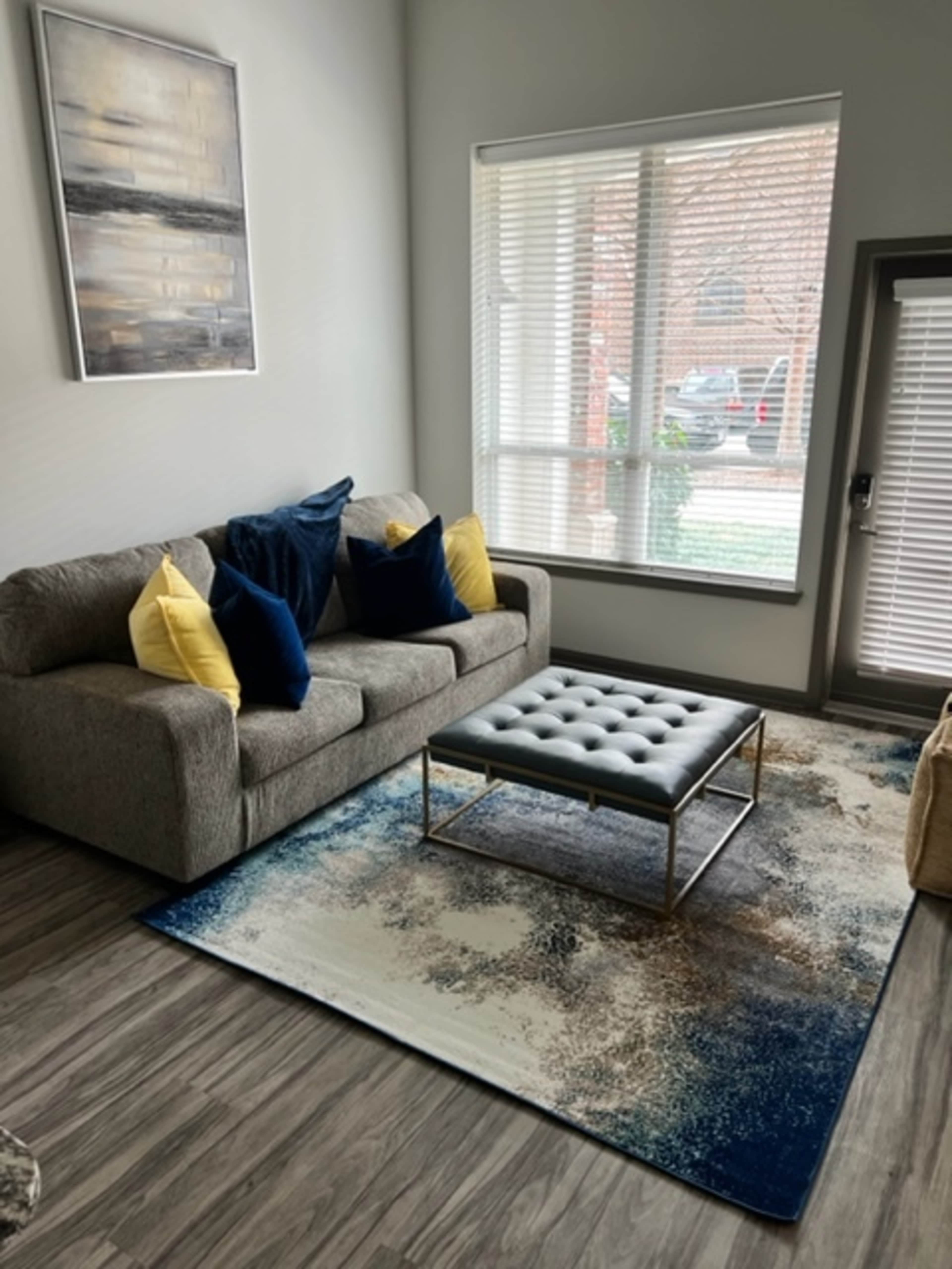 The image shows a living room with a grey sofa, two yellow and blue pillows, a tufted ottoman on a patterned rug, and large windows allowing natural light.