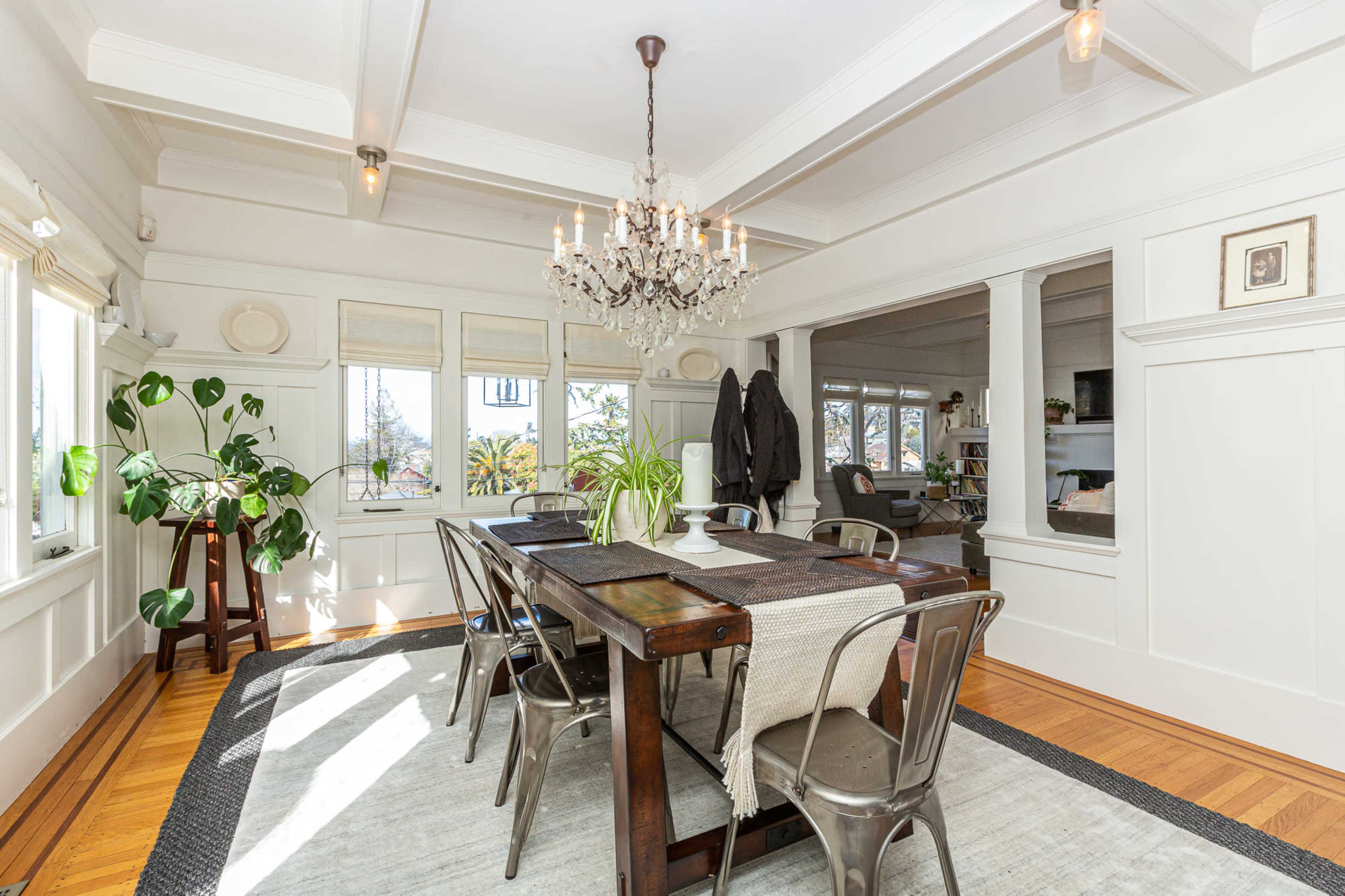 A dining room with a wooden table, metal chairs, a chandelier, and large windows letting in natural light, surrounded by a neutral color palette.