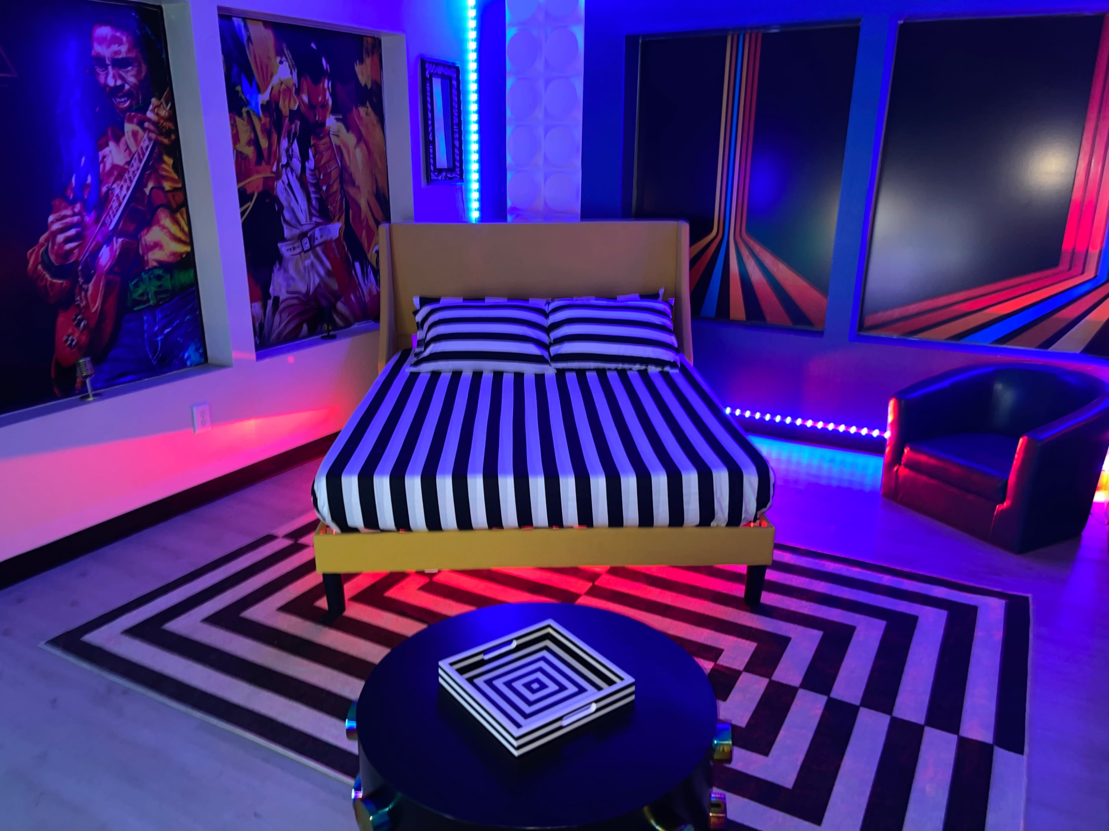 The scene depicts a bedroom with a yellow bed featuring black and white striped bedding, surrounded by colorful wall artwork and vibrant LED lights.