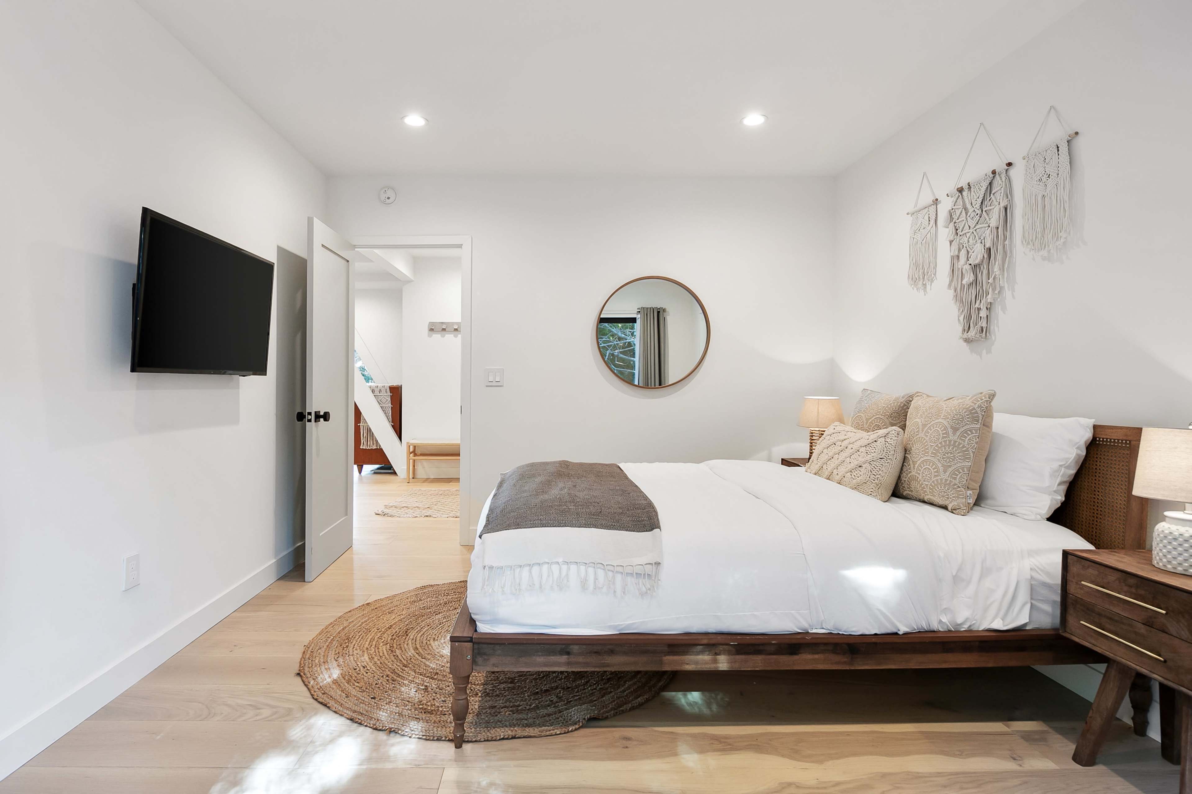 The image shows a modern bedroom featuring a bed with white linens, a round mirror on the wall, wooden furniture, and soft lighting.