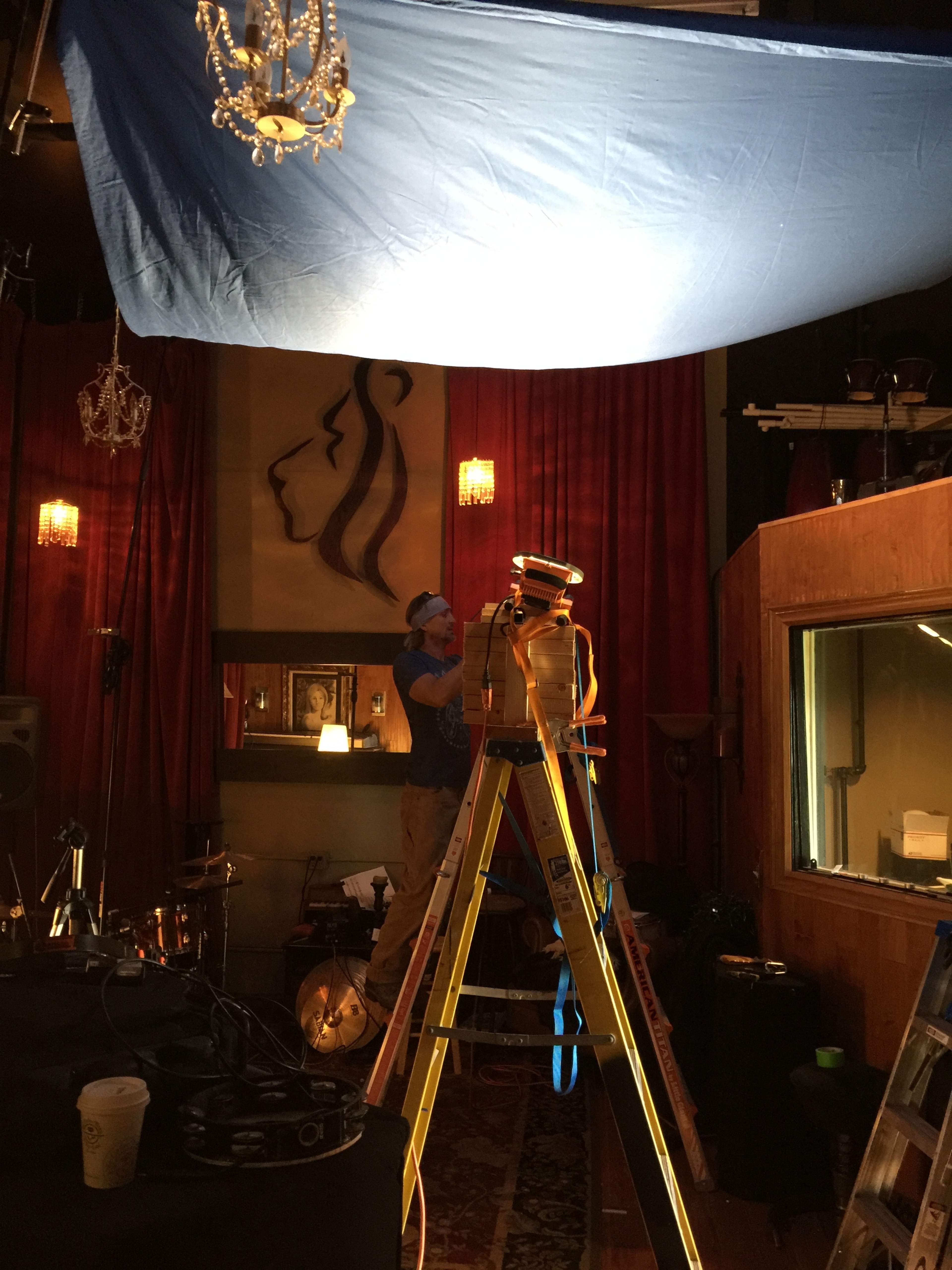 A man adjusts a blue fabric draped overhead in a recording studio with musical instruments and decorative lighting in the background.