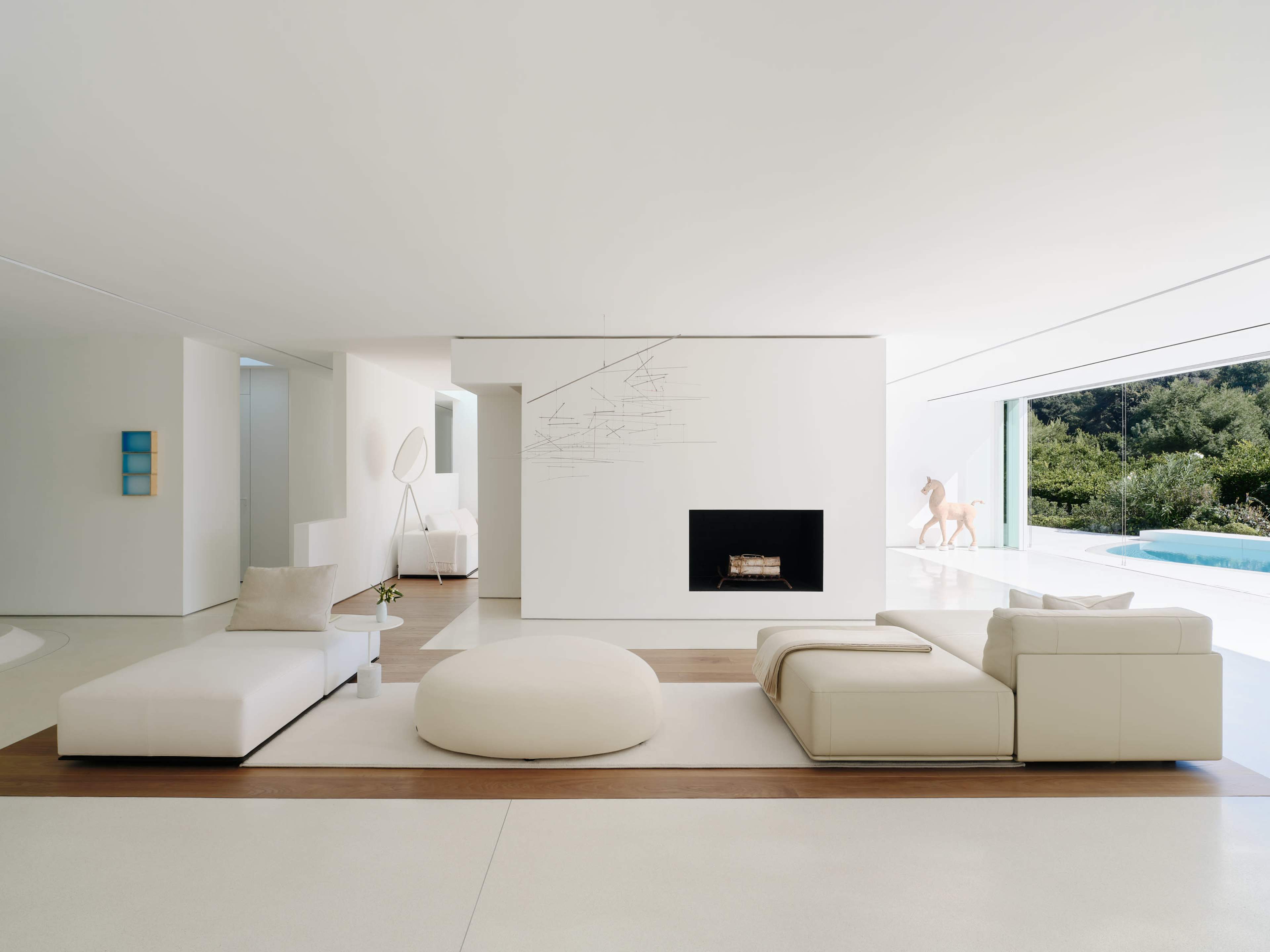 The image showcases a modern living room with a white sectional sofa, a circular ottoman, a minimalist fireplace, and large windows overlooking a pool and greenery.