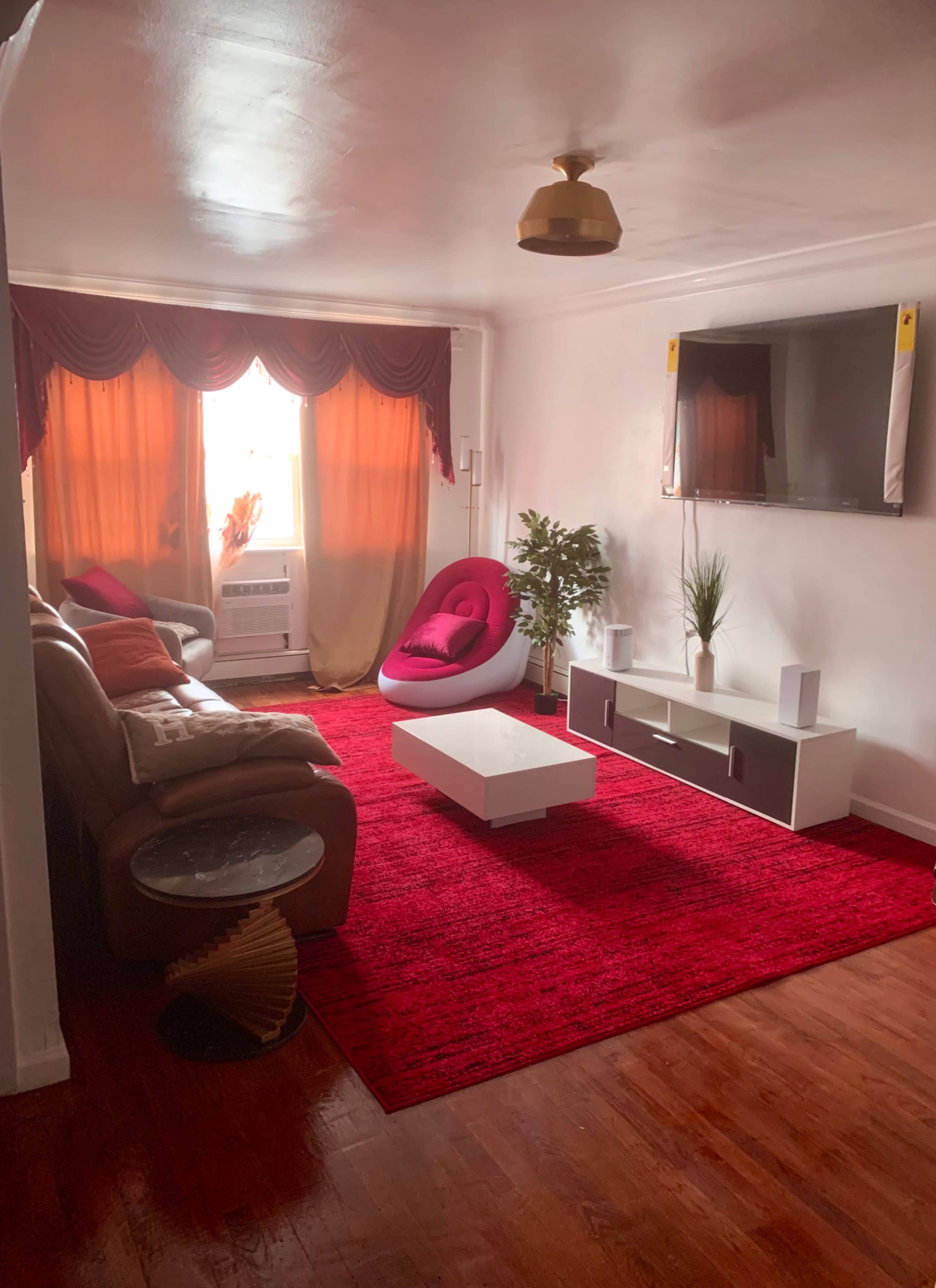The living room features a red area rug, a white television stand, a large window with curtains, and a mix of seating including a brown couch and a modern white chair.
