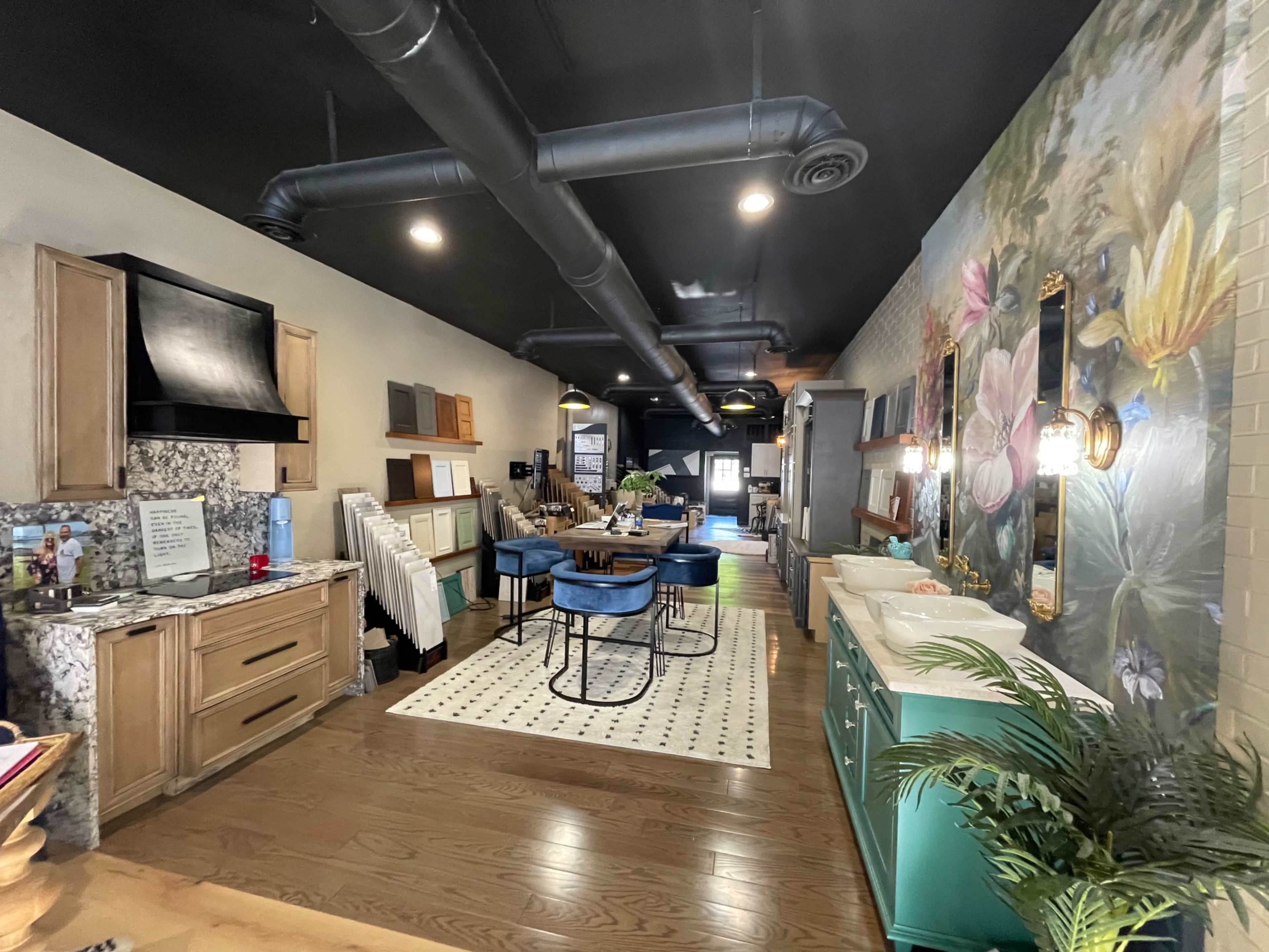 A modern kitchen and dining area with wooden cabinetry, a large table surrounded by blue chairs, and a wall adorned with a floral mural.