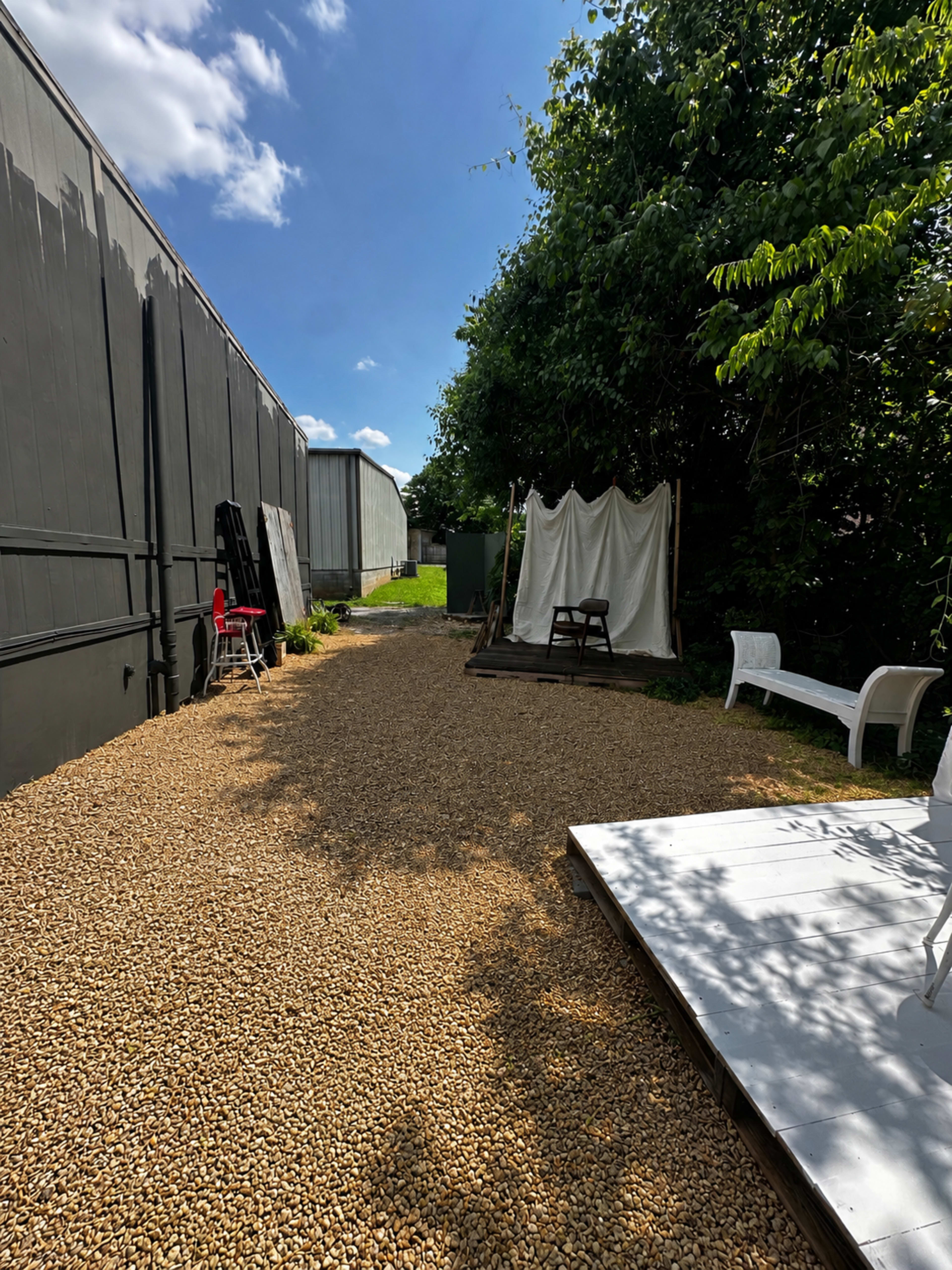The scene shows a gravel pathway alongside a building, flanked by trees and a white structure with a draped fabric.