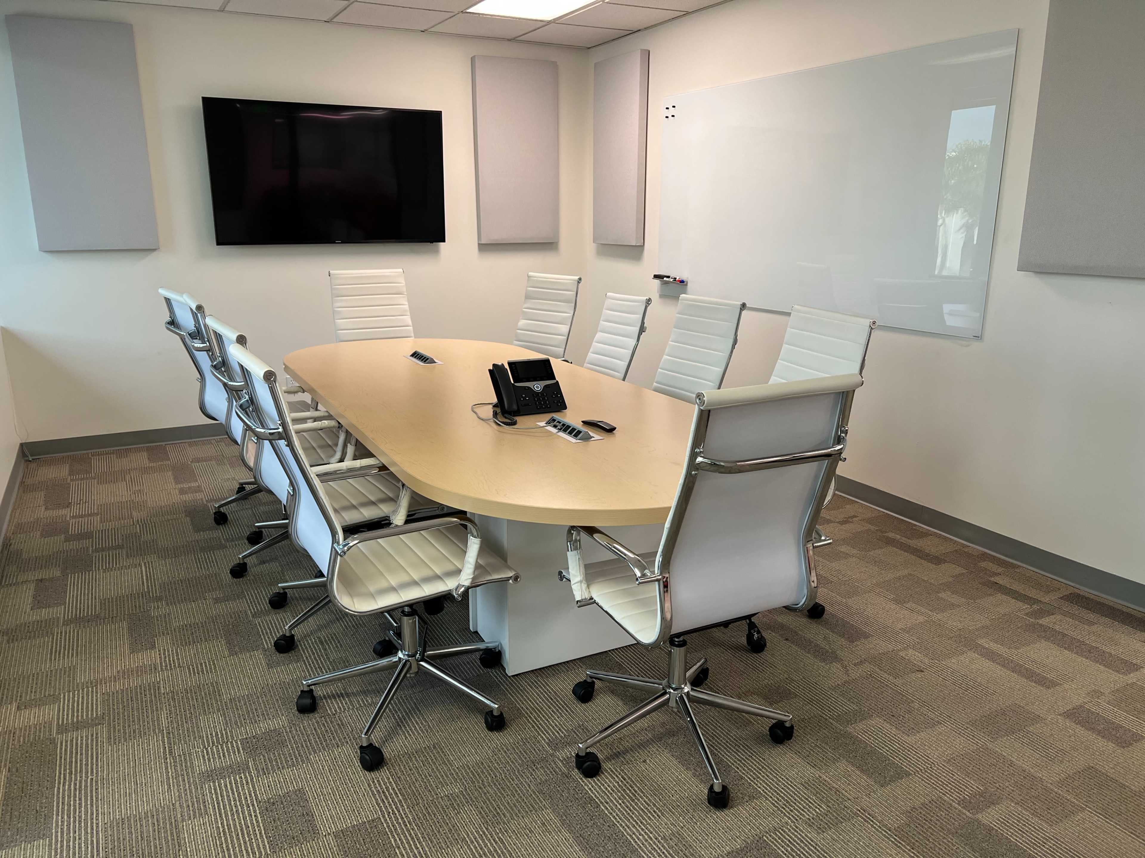 A modern conference room is set up with an oval table surrounded by white ergonomic chairs, a wall-mounted screen, and a whiteboard.