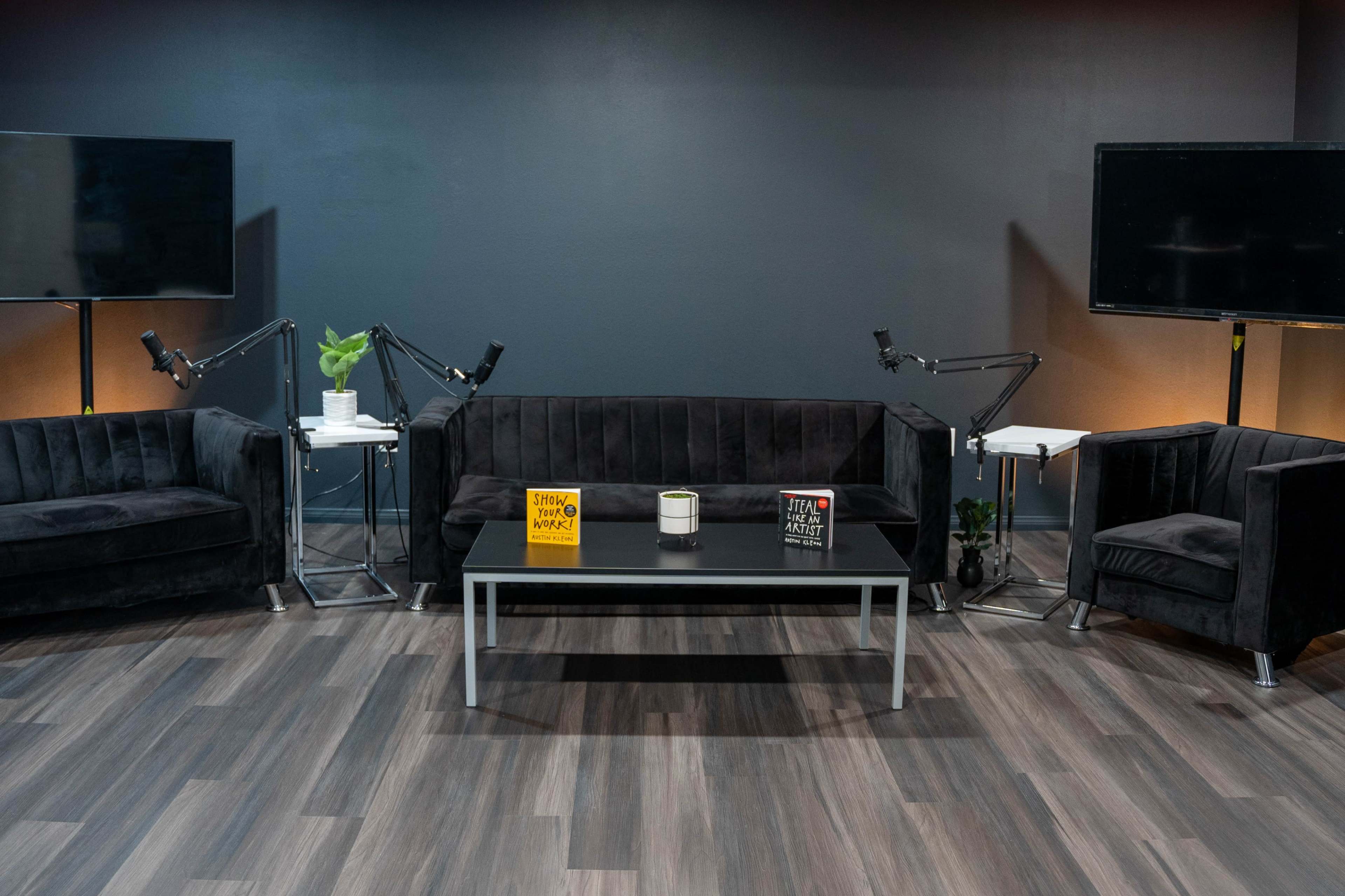 The scene features a modern seating area with two black sofas and a chair arranged around a central table, flanked by two large monitors.