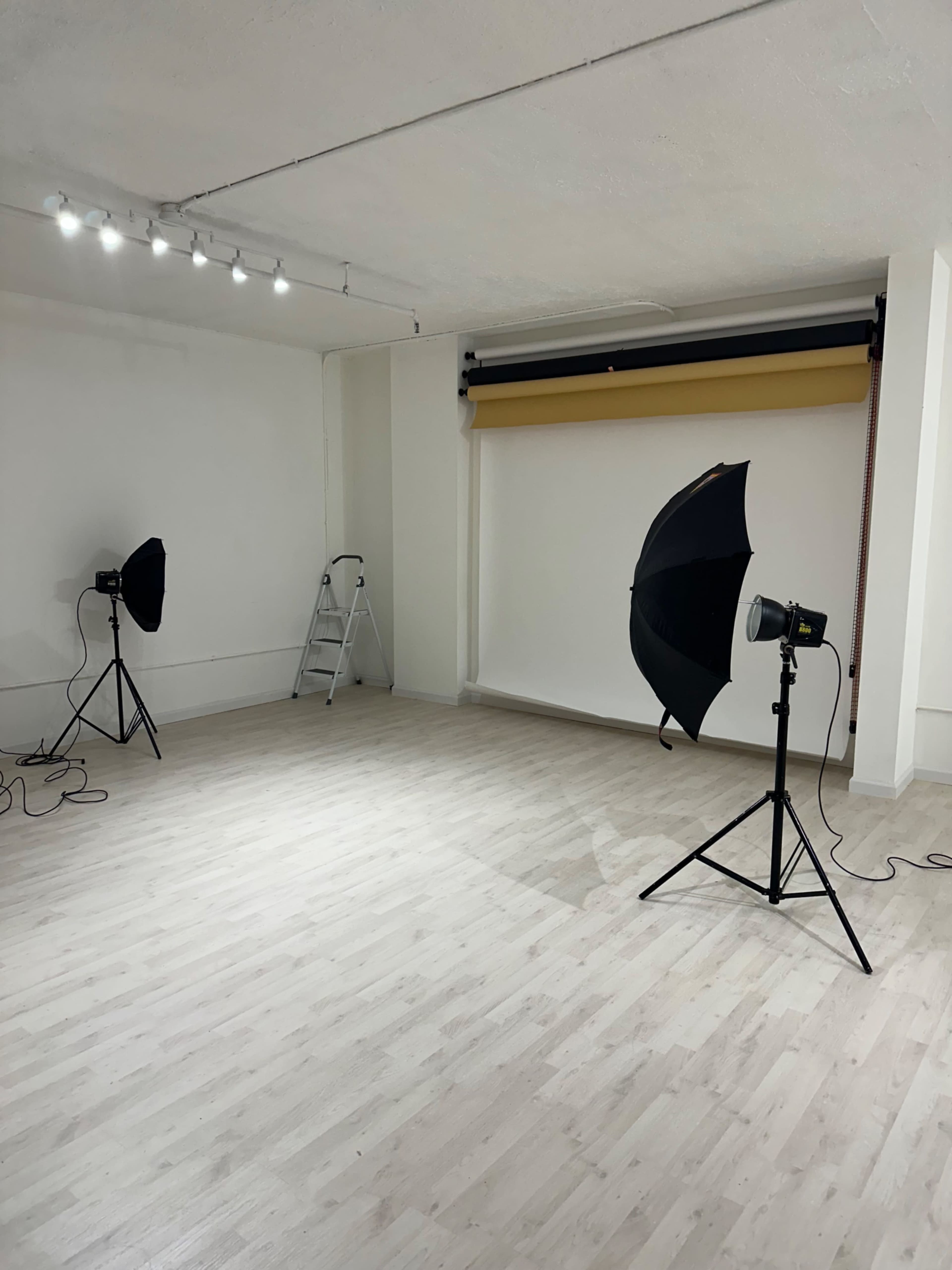 The image shows a well-lit photography studio with two softbox lights, a ladder, and a backdrop setup against a plain white wall.