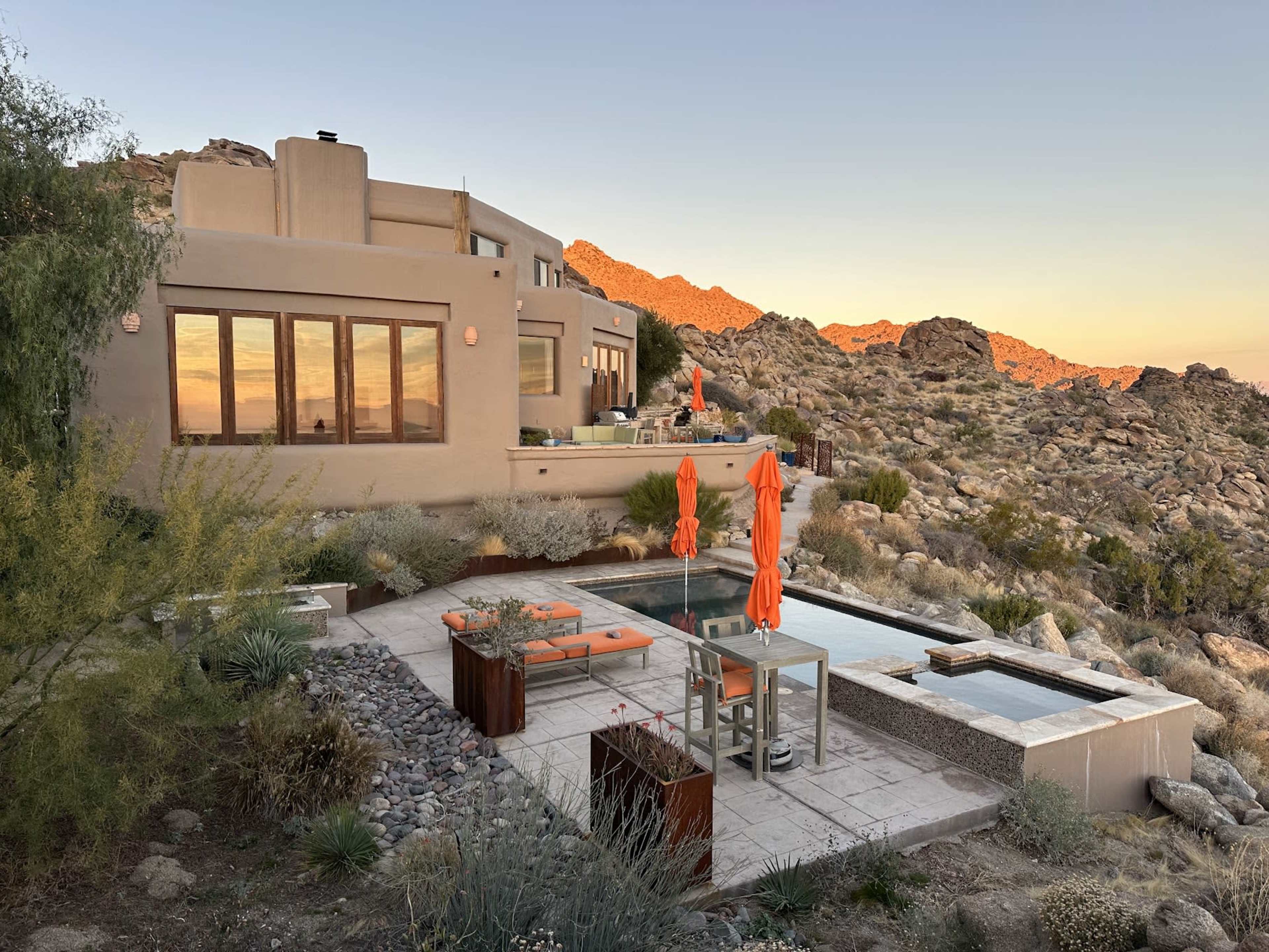 A modern desert house with large windows overlooks rocky terrain and features a swimming pool surrounded by orange lounge chairs and umbrellas.