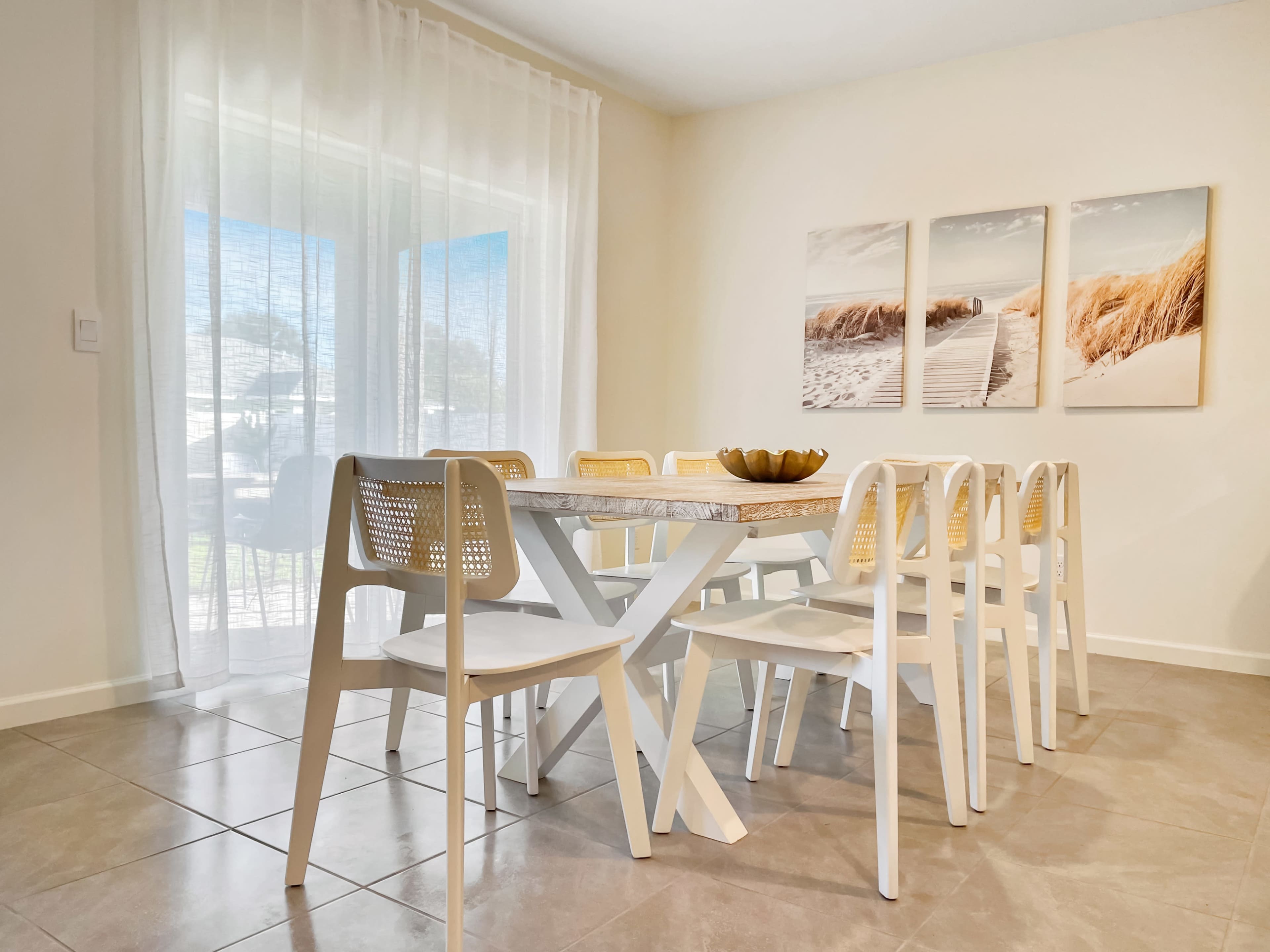 A modern dining area features a white table surrounded by eight chairs, with large windows and beach-themed artwork on the walls.