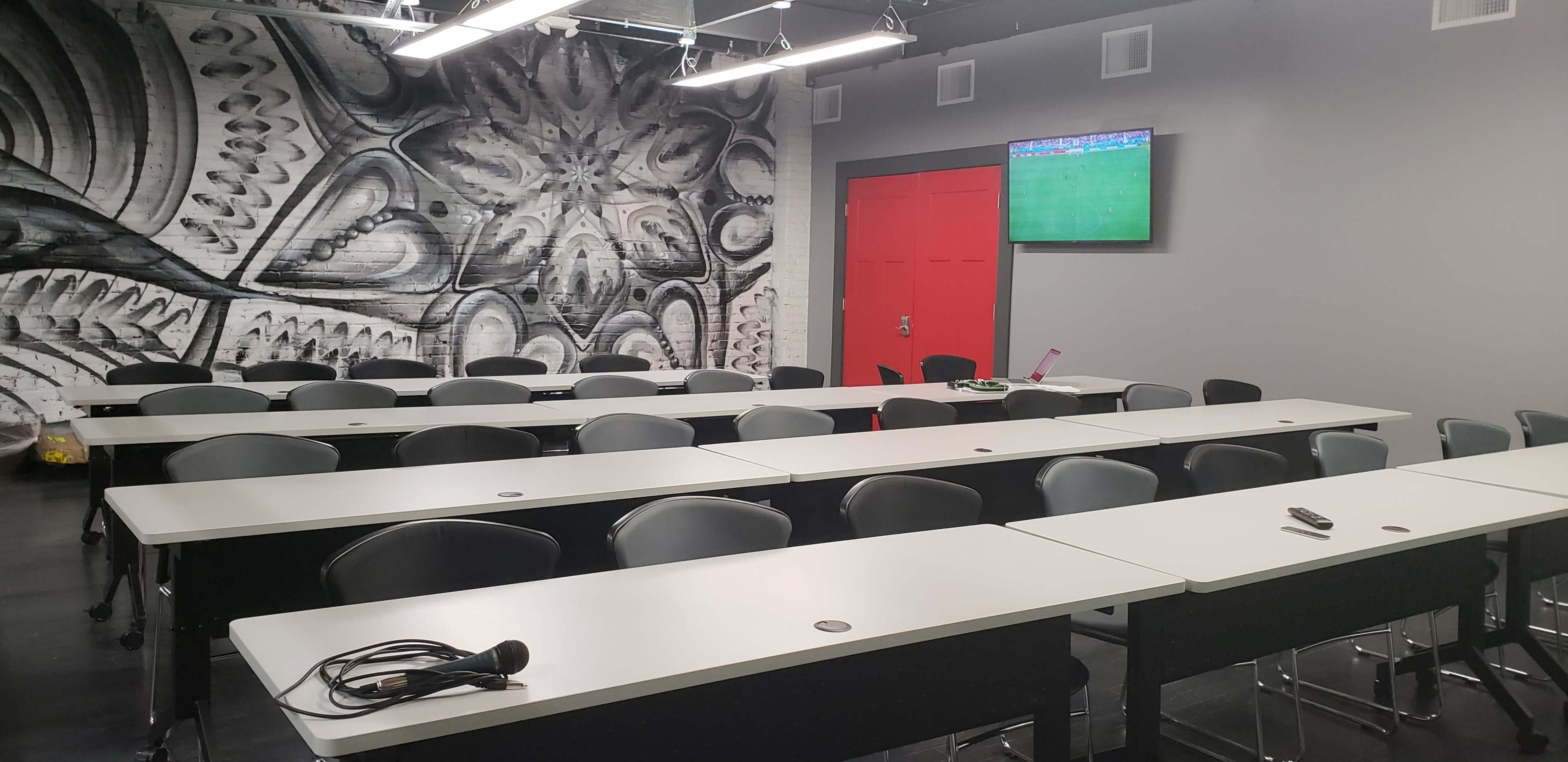 The image shows a modern classroom with rows of tables and chairs, a large mural on the wall, and a television screen displaying content in the corner.