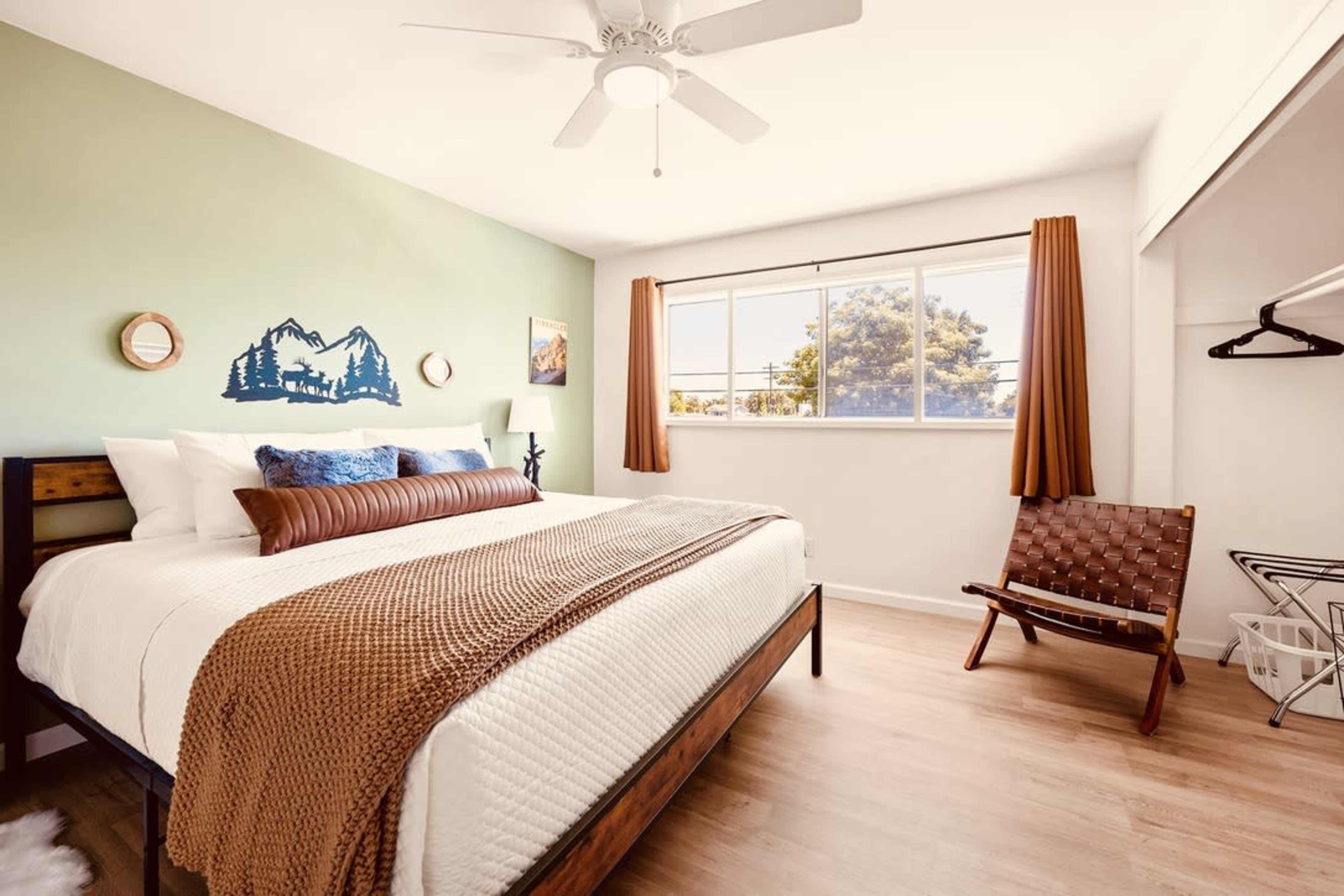 A well-lit bedroom features a large bed with a brown throw blanket, a wall-mounted mountain mural, and a window with orange curtains overlooking a view.