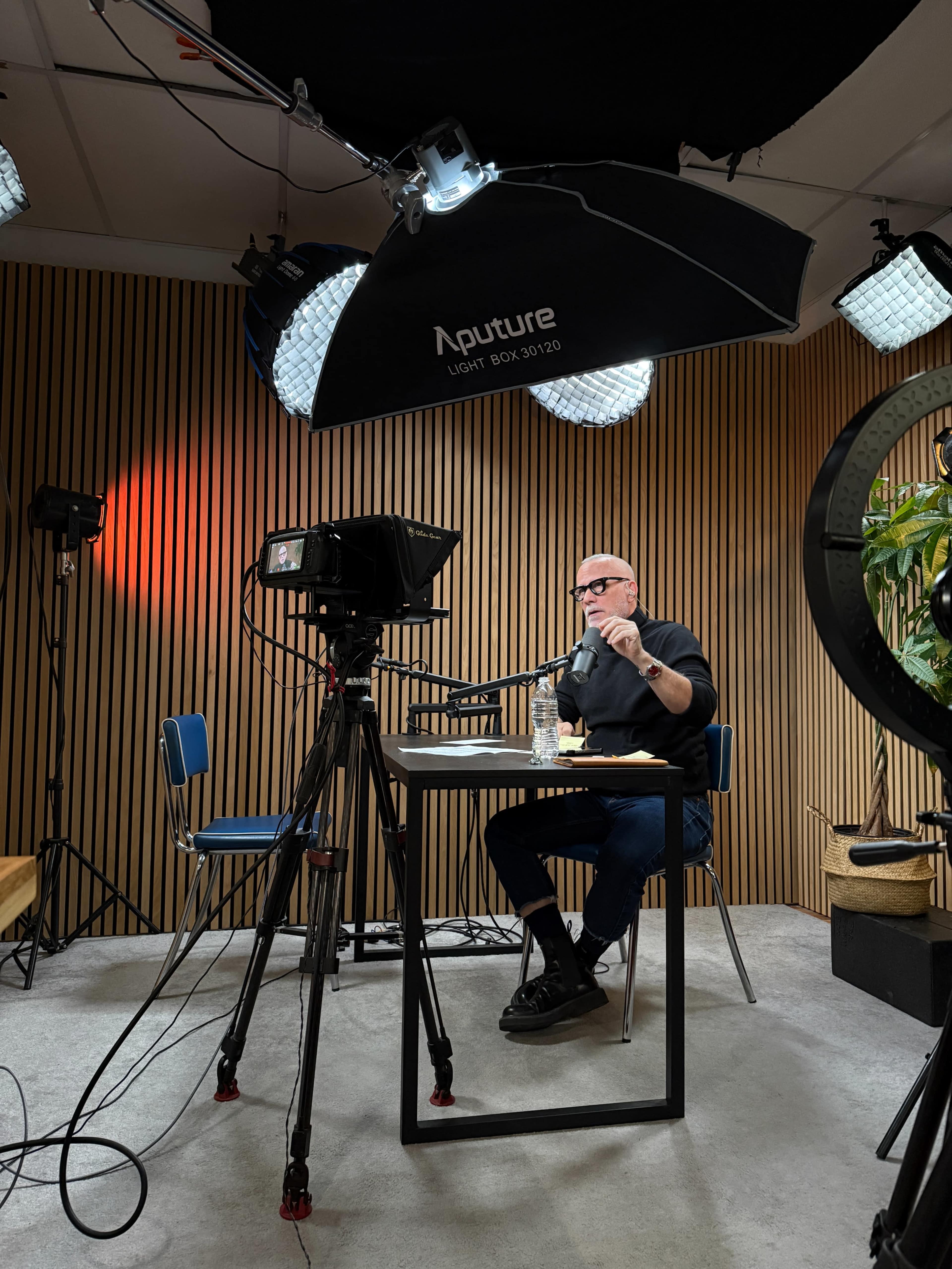 A person wearing glasses sits at a table in a well-lit studio, holding a microphone while surrounded by cameras and studio lighting.