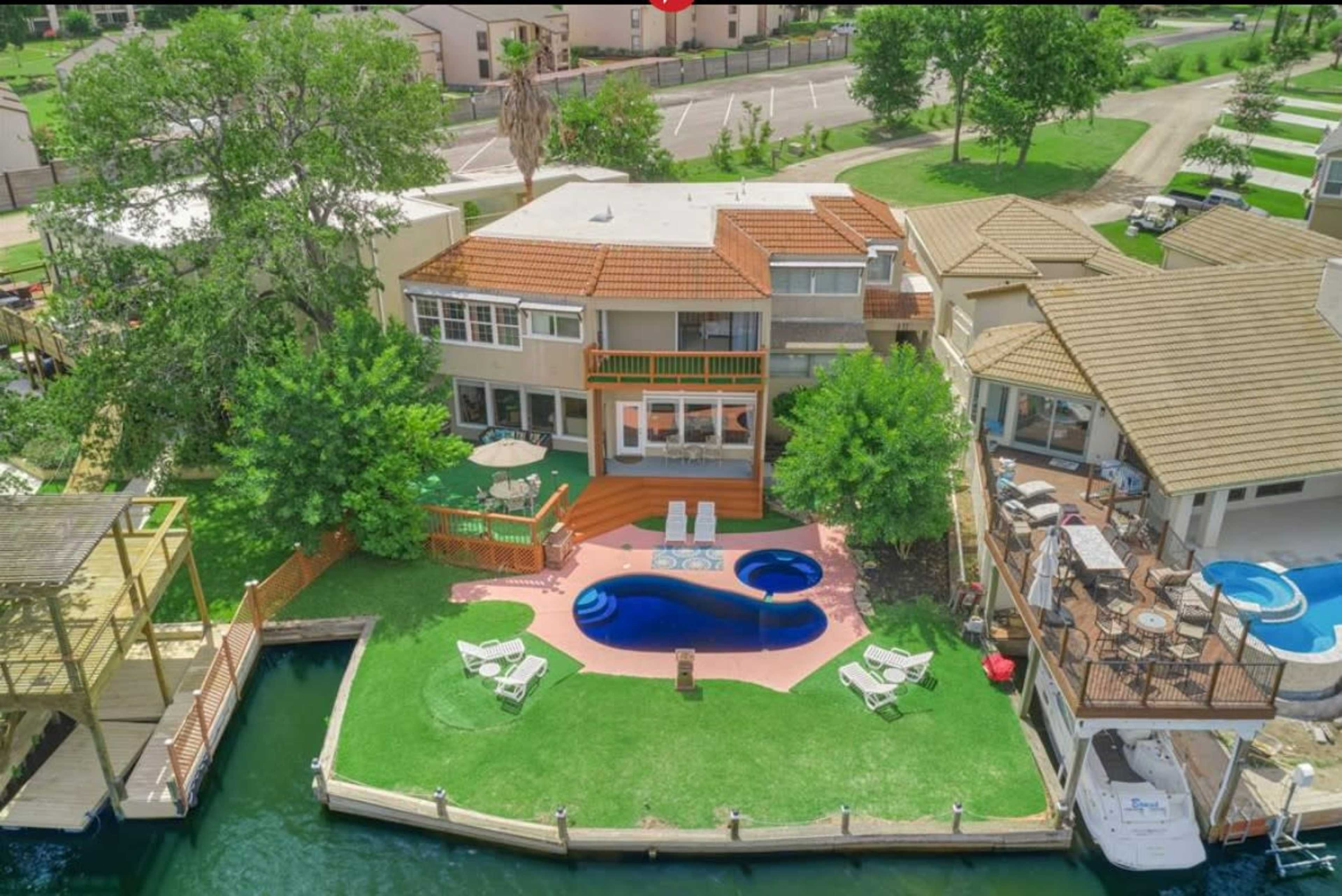 The image shows a large waterfront property with a swimming pool, multiple decks, and boats docked on a green lawn alongside a canal.