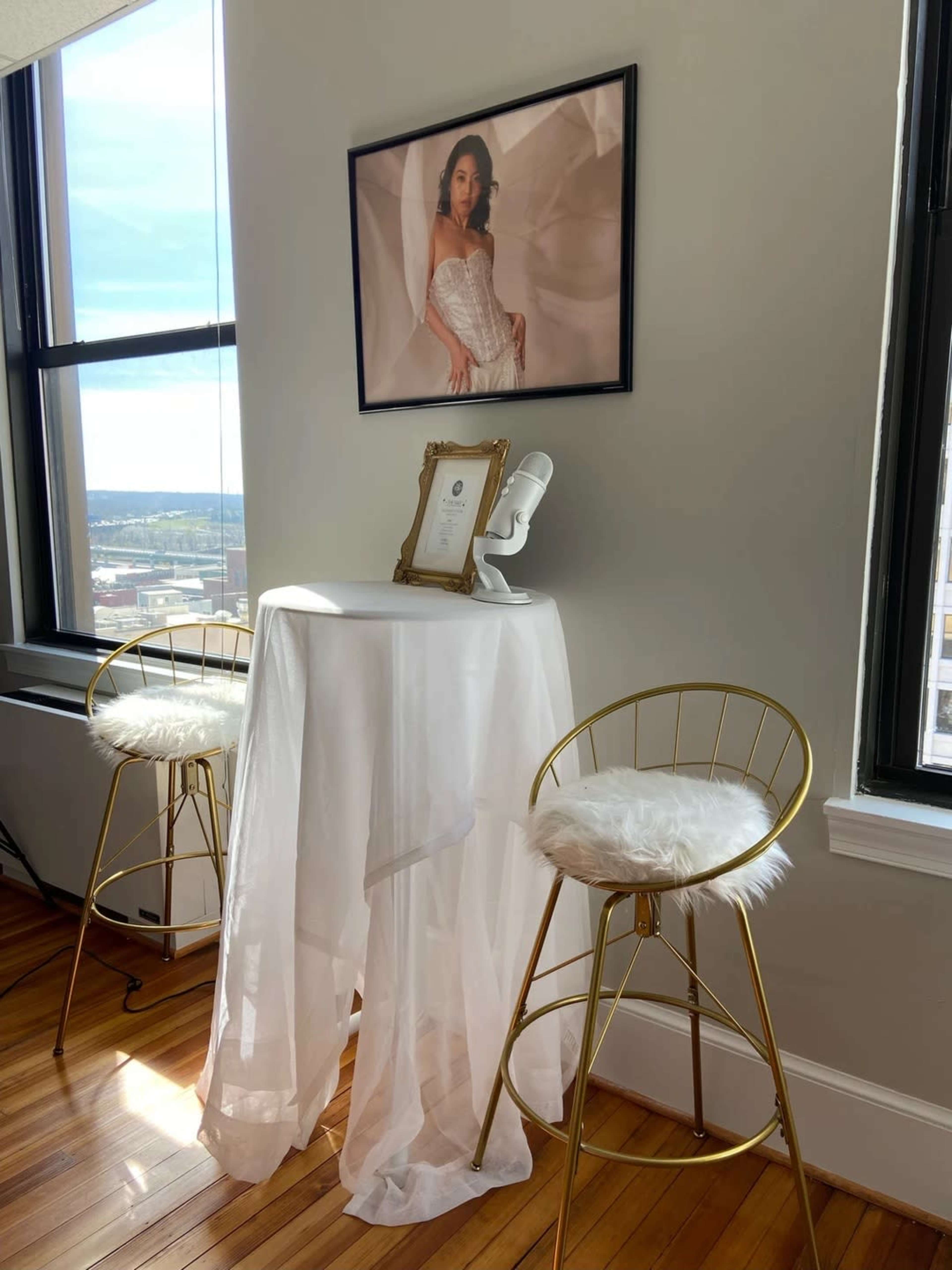 A small table draped with white fabric and adorned with a framed photo and a decorative bust stands between two gold bar stools in a bright room with large windows.