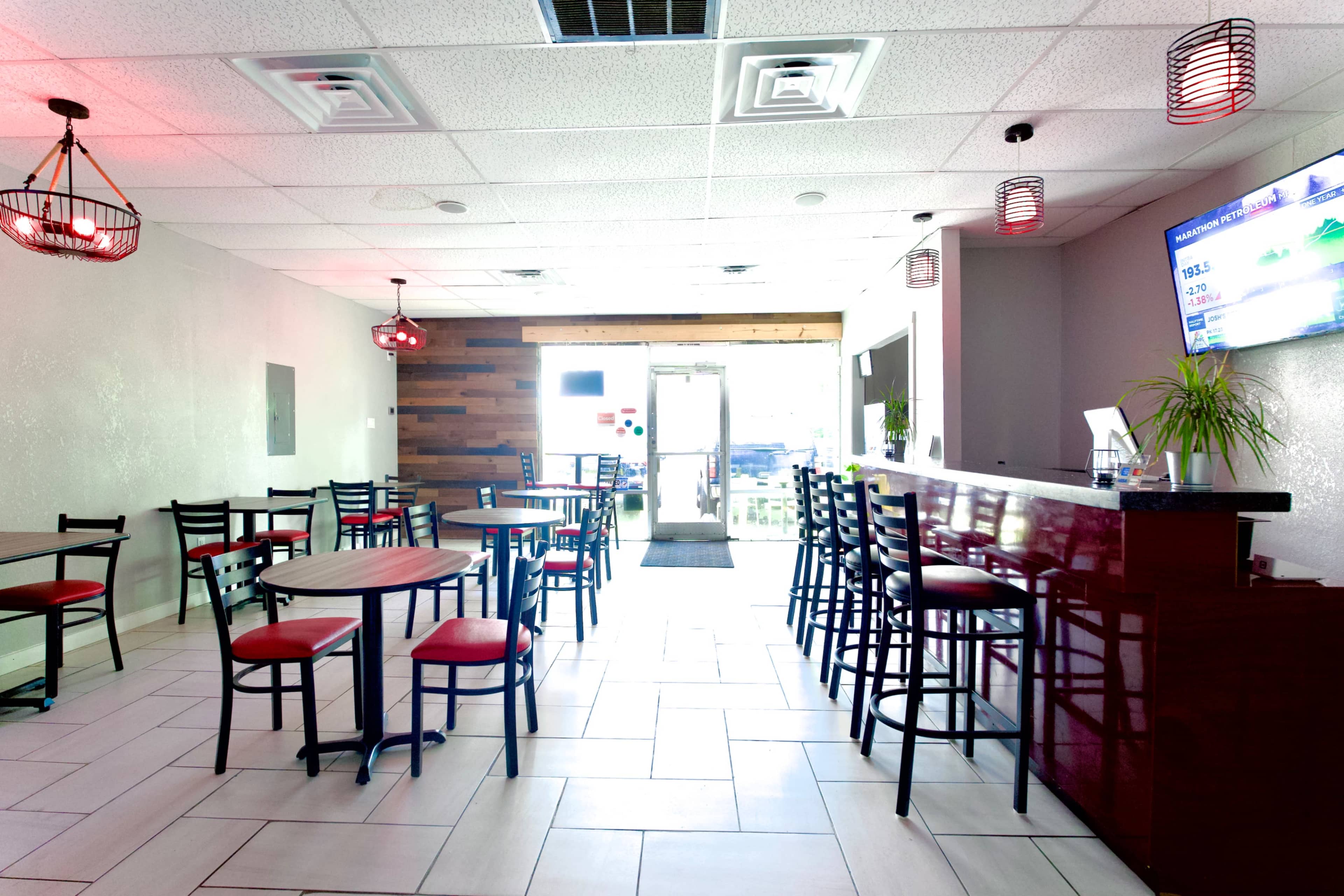 The image shows the interior of a restaurant or café featuring tables and chairs, a bar area, and large windows that provide natural light.
