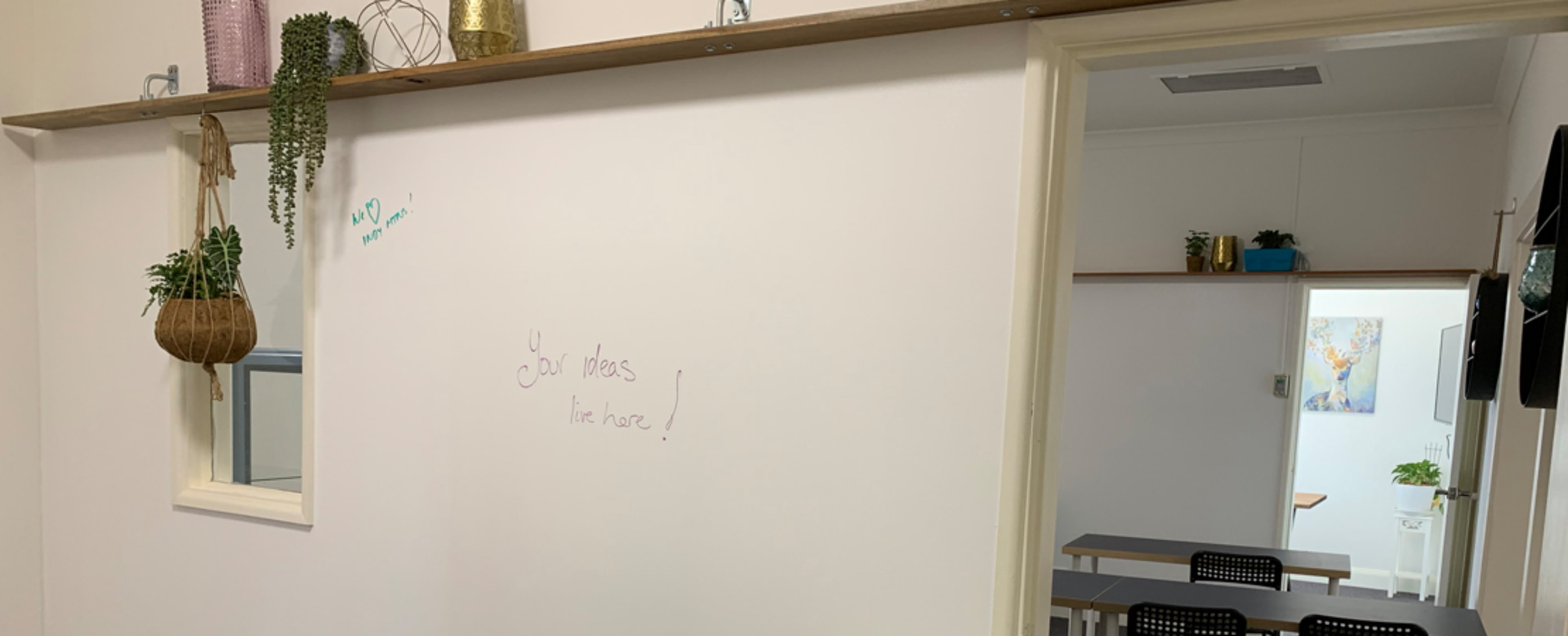 The image shows a light-colored wall with a hanging shelf adorned with plants, and a doorway leading to another room with a table and chairs.