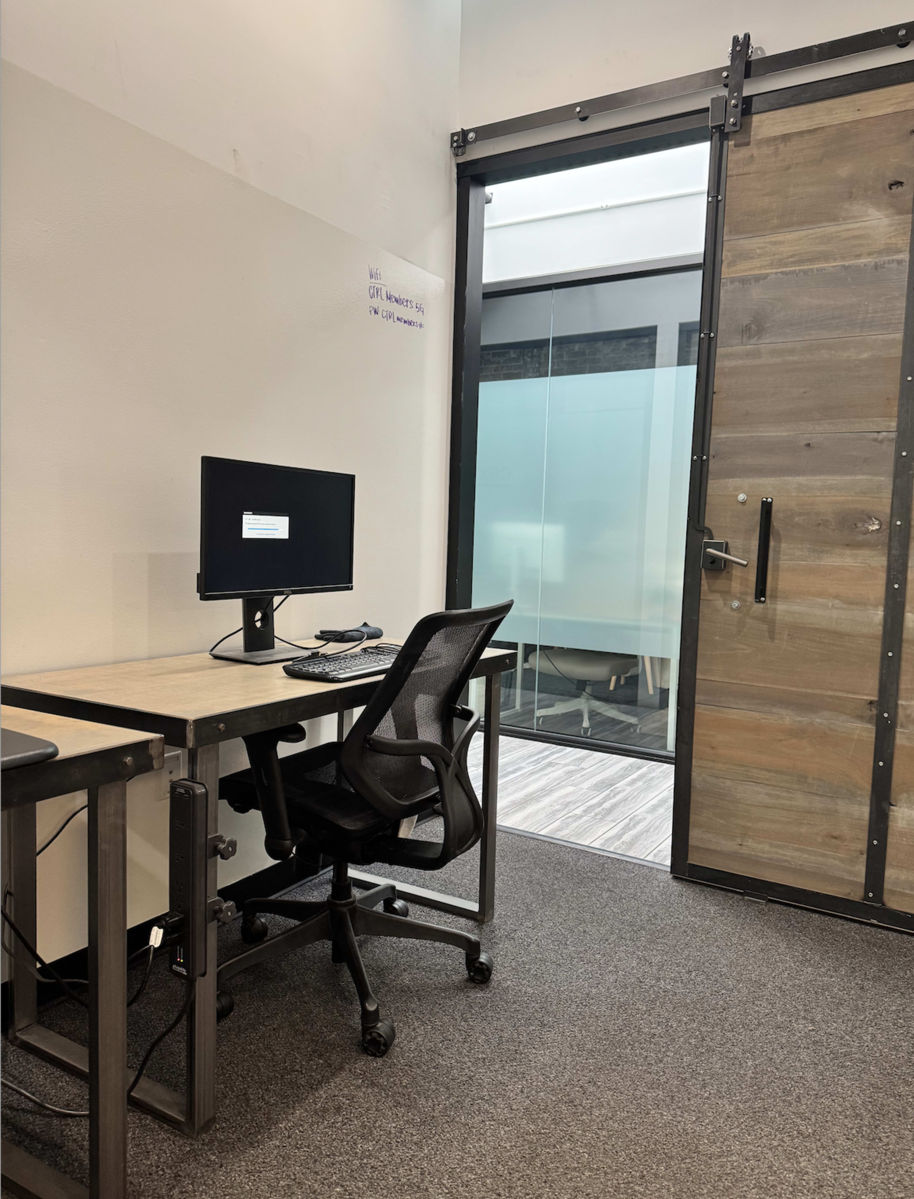 The image shows a workspace featuring a desk with a computer monitor and an office chair, adjacent to a sliding glass door leading to a meeting room.