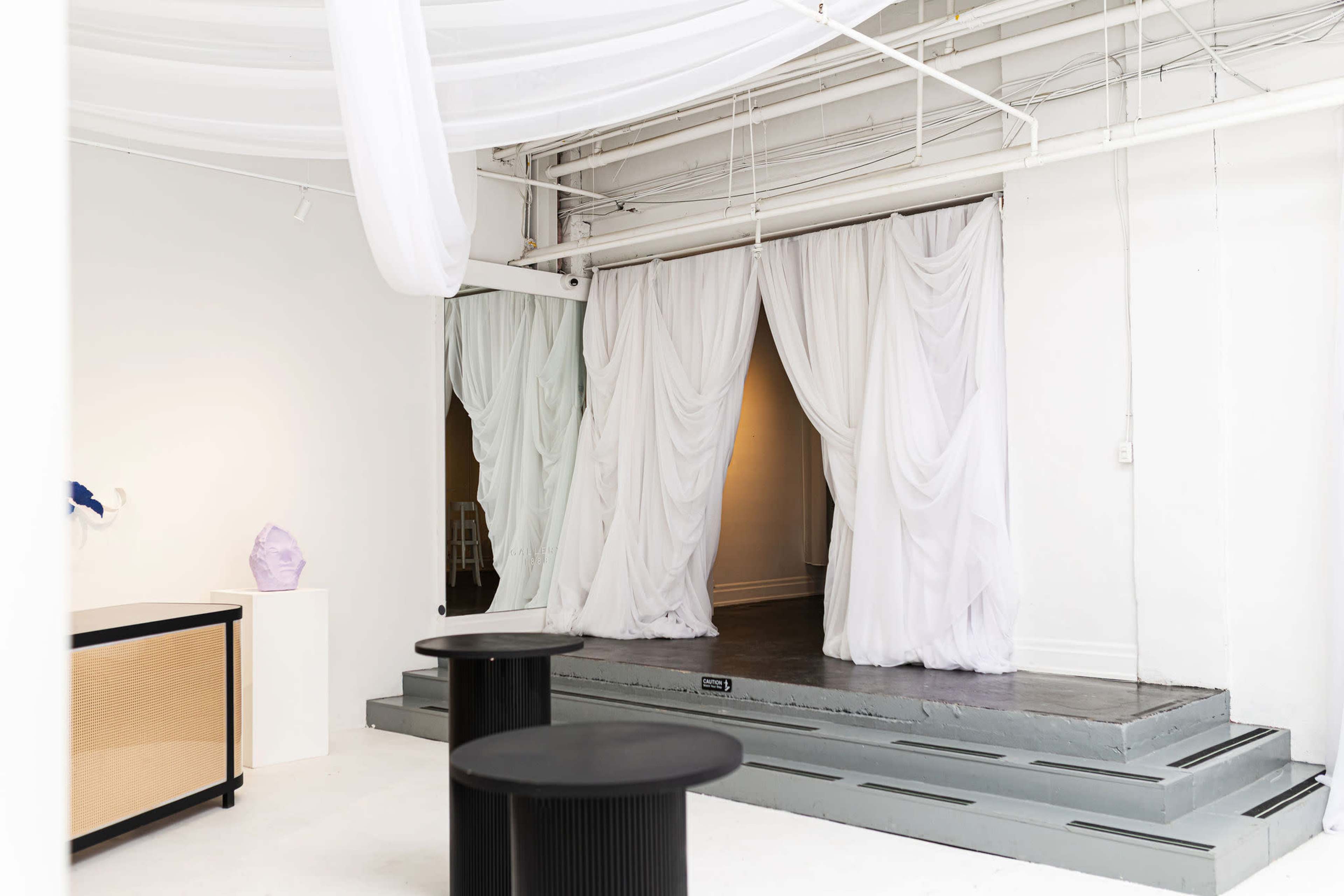 The image shows a minimalist interior space featuring white draped curtains, a small stage area, and two black circular tables.